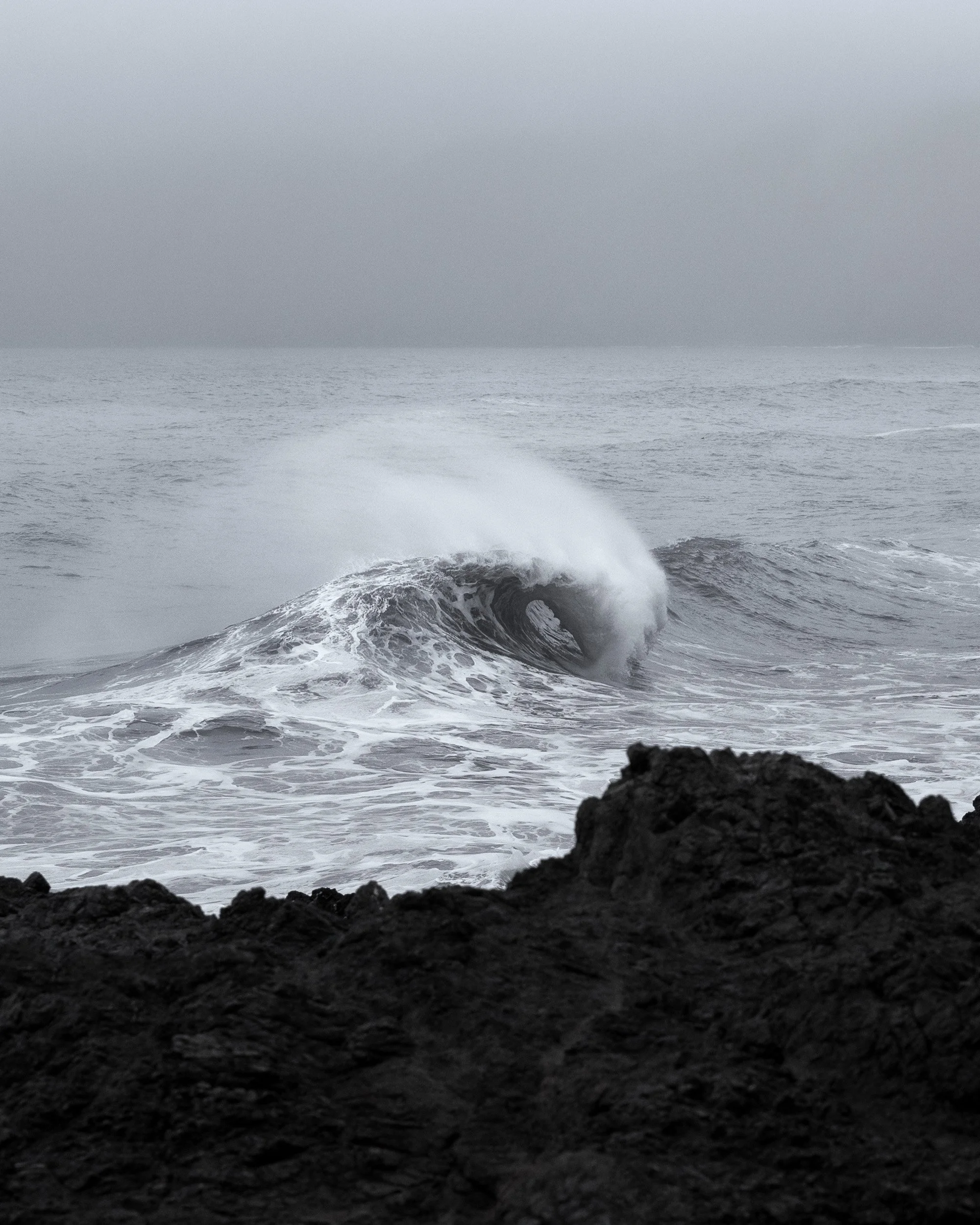 A wave curls and breaks in heavy grey seas beyond dark rocks in this black and white seascape.
