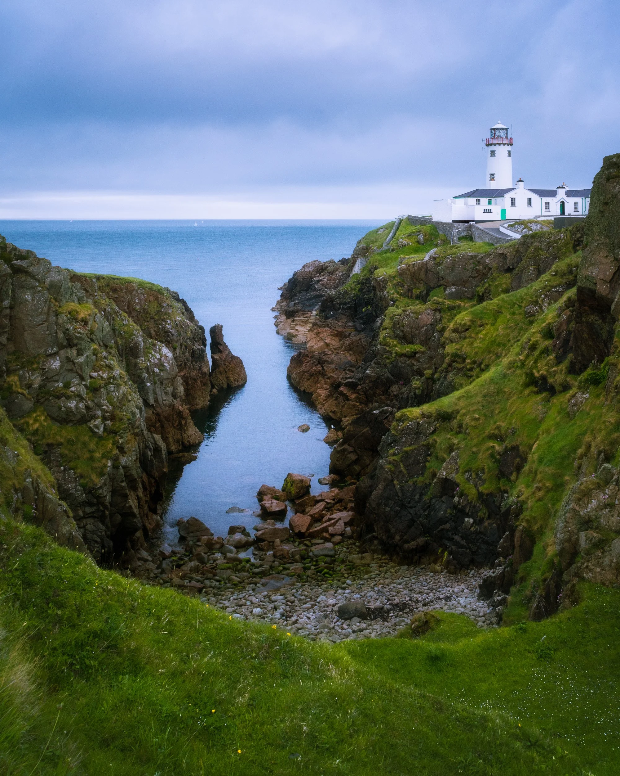 Fanad Head Lighthouse stands white against a grey sky above a rocky sea inlet on the Donegal coast, Ireland.