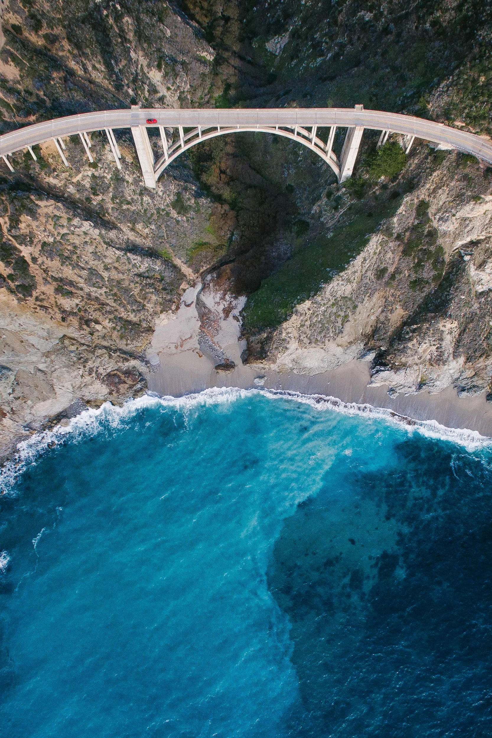 Aerial view looking straight down on Bixby Creek Bridge and a red car crossing it, with turquoise surf breaking on the Big Sur coast below.
