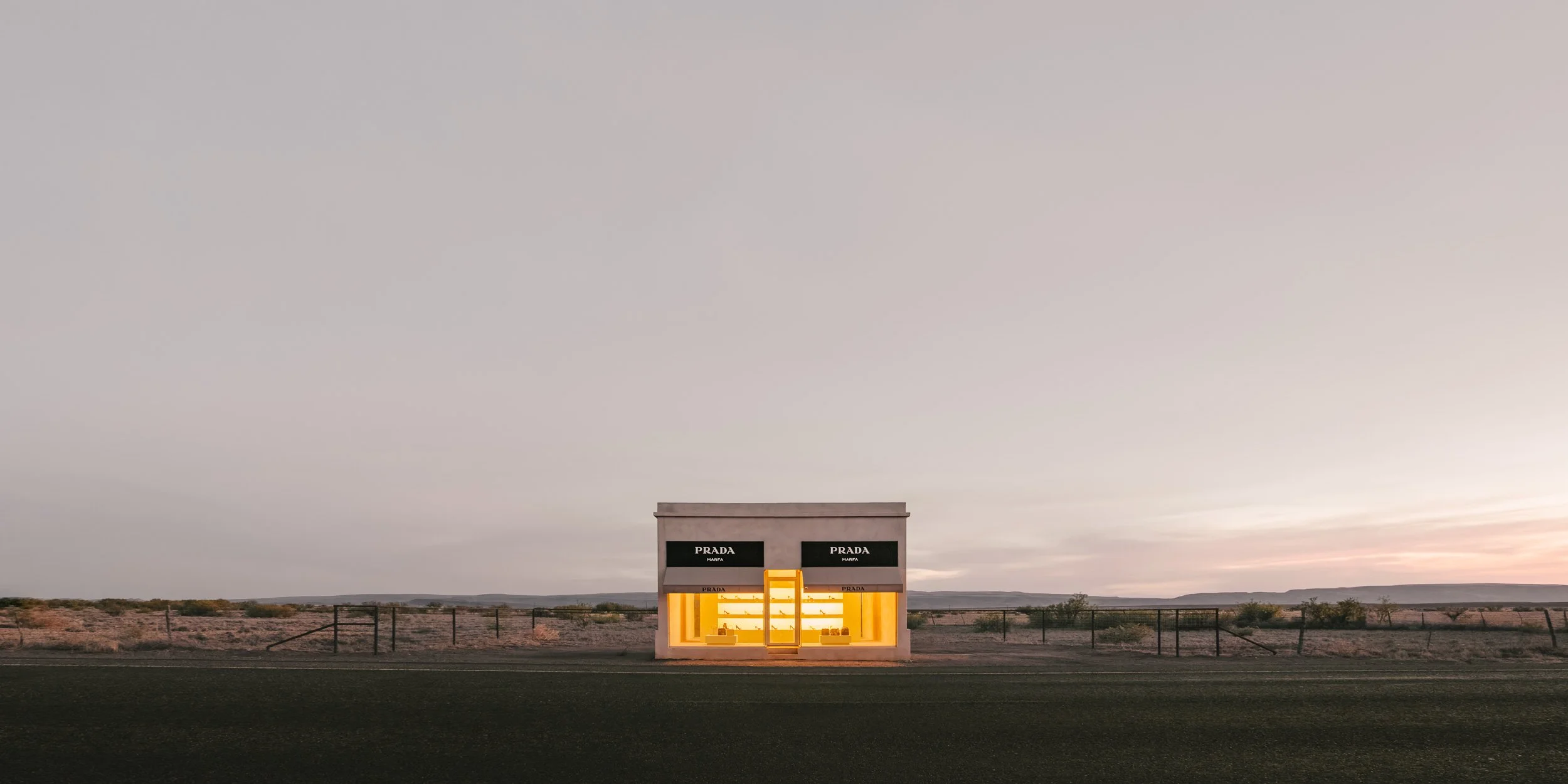 The Prada Marfa art installation glows warm yellow at dusk against a pale West Texas sky near Valentine, Texas.