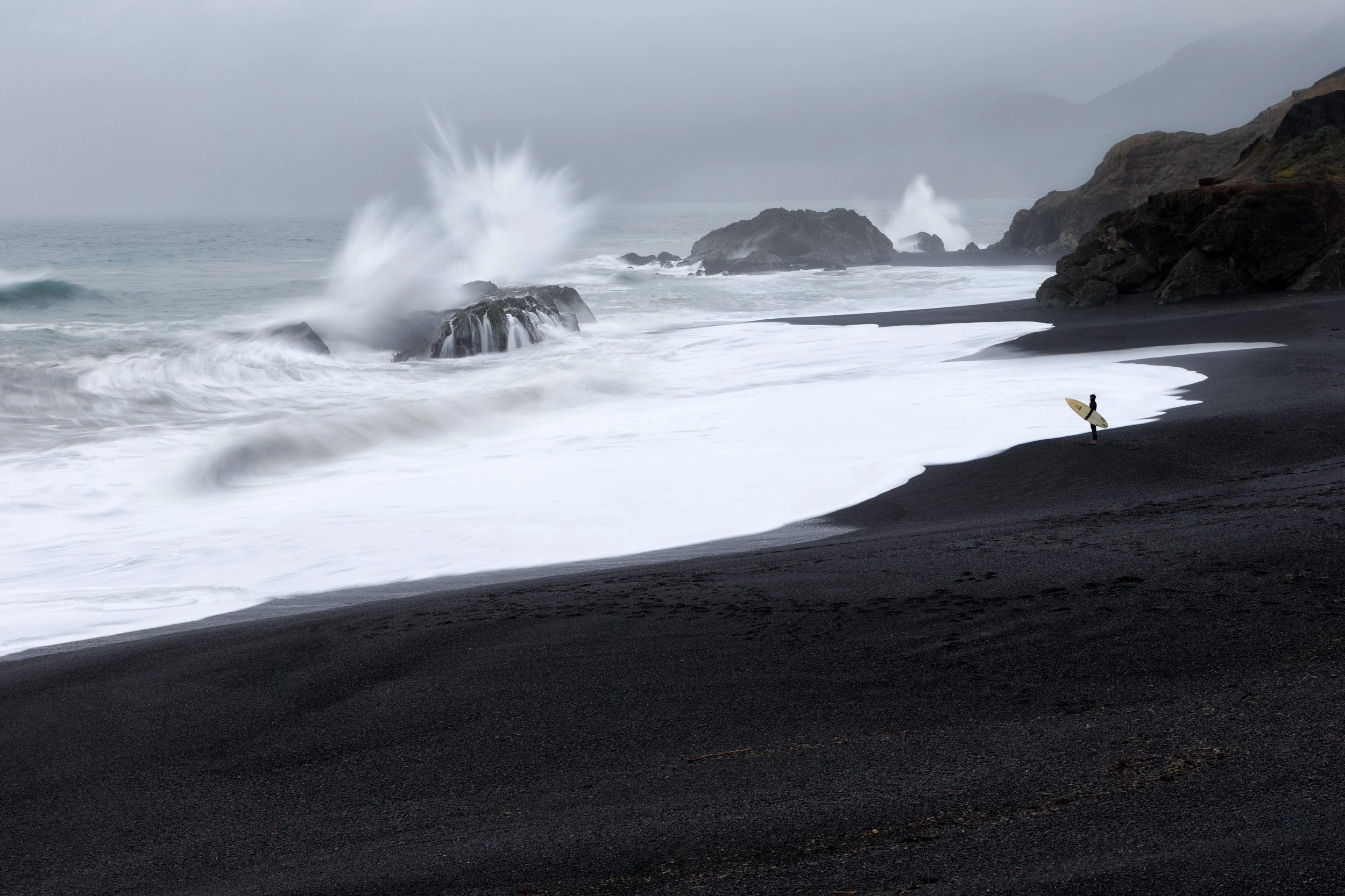 A lone surfer holding a board watches storm waves explode against sea stacks from a black sand beach in Shelter Cove, California