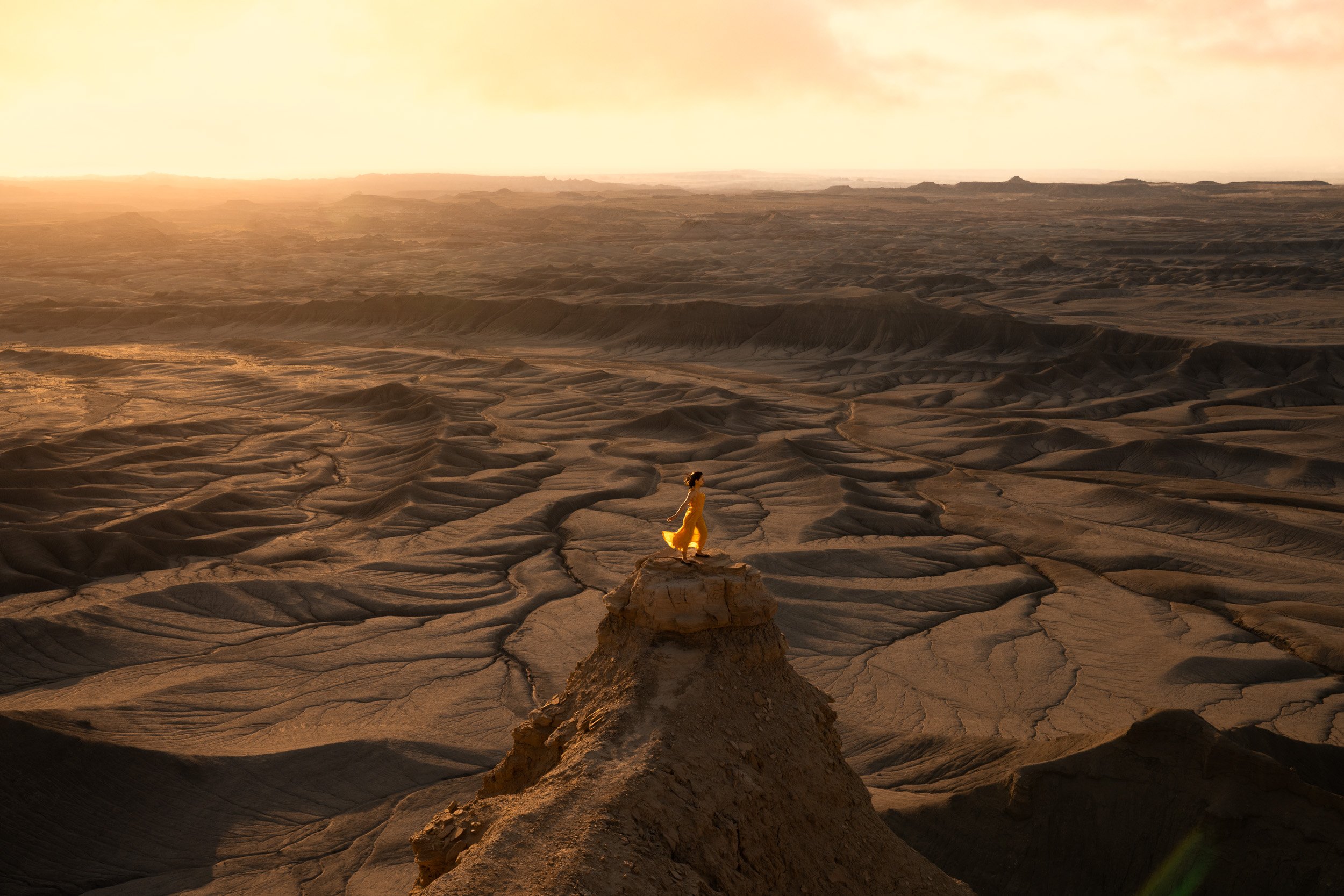 A woman in a yellow dress stands on a rock pedestal above endless grey badlands at Moonscape Overlook near Hanksville, Utah.