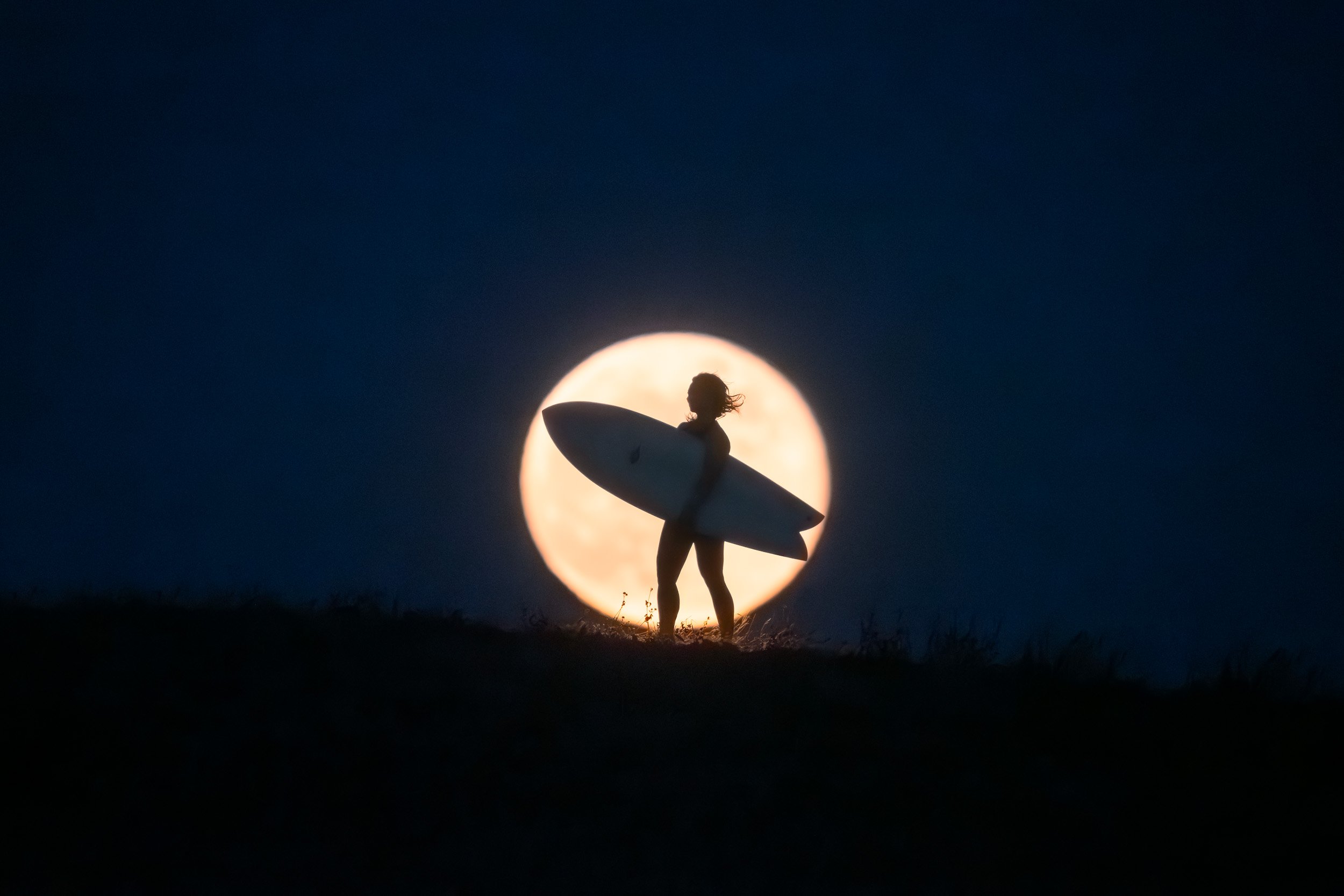 A surfer carrying a board stands silhouetted against a rising full moon on a dark ridgeline.
