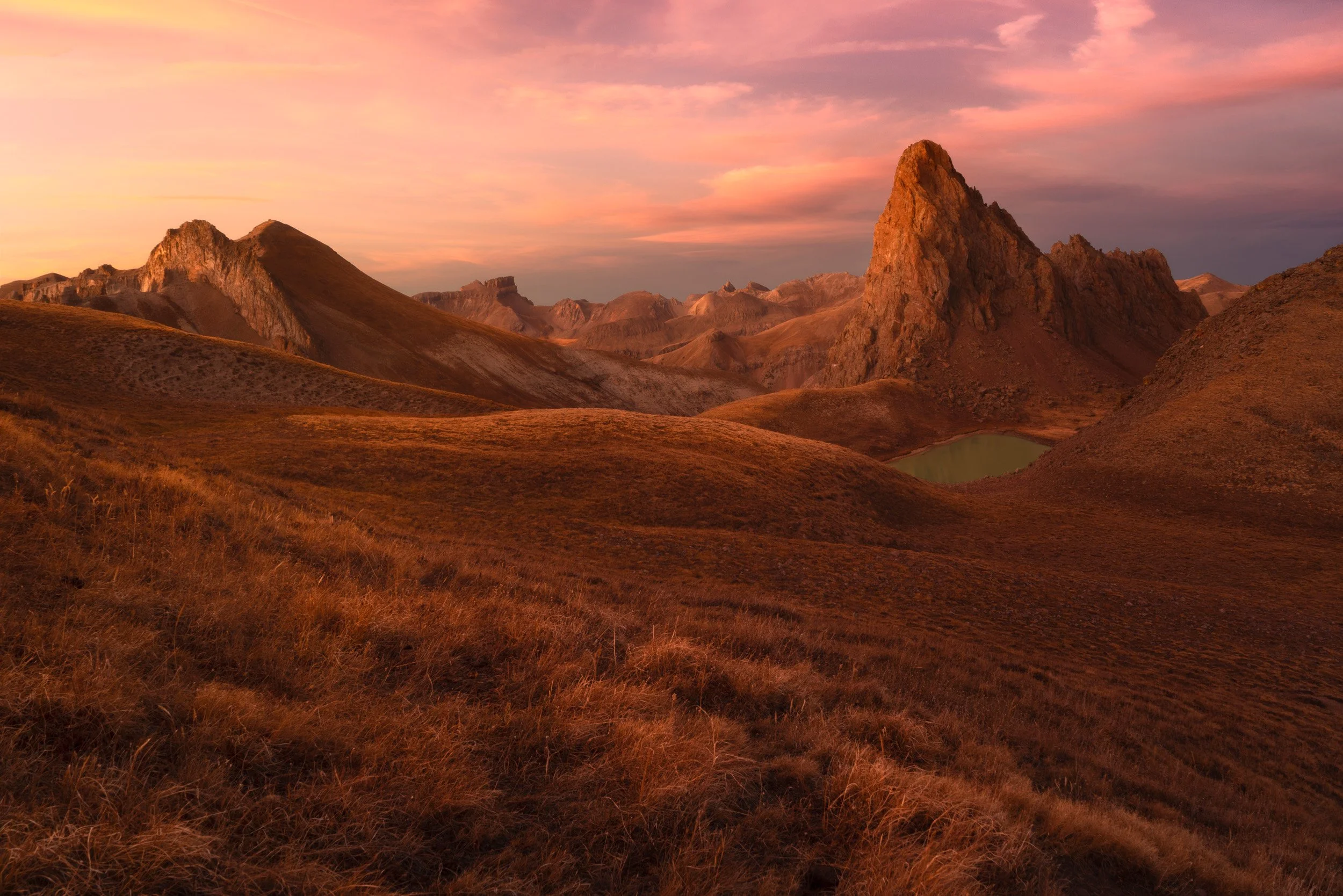 Pink sunset light washes over Dragon's Back and a small alpine tarn in a Colorado grassy basin.