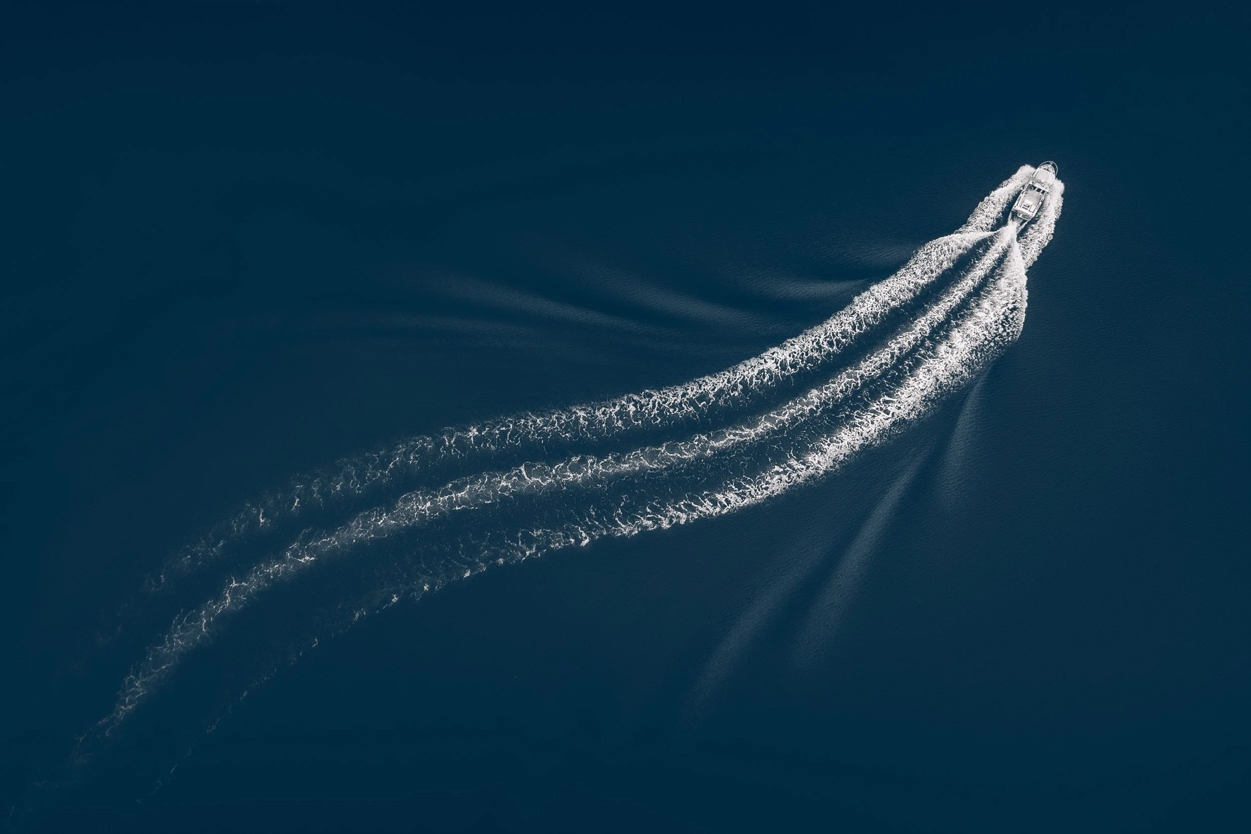 Aerial view of a small boat carving a long white wake across dark ink-blue water, Alaska.