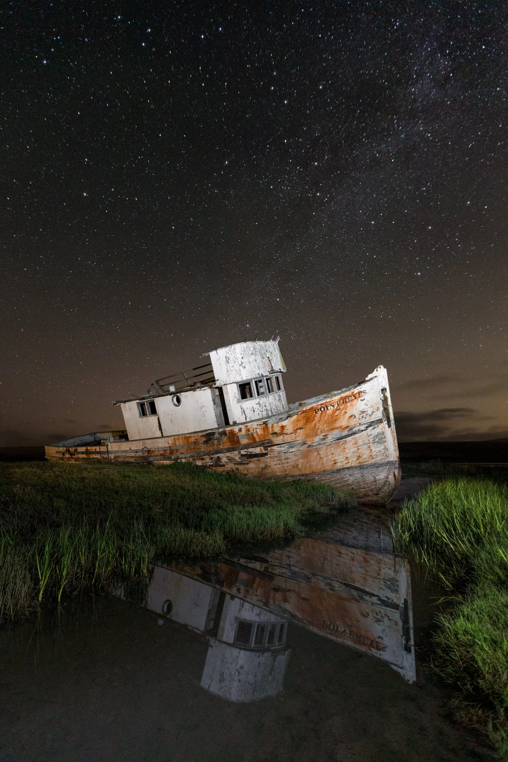 The weathered Point Reyes shipwreck rests in marsh grass beneath a star-filled night sky, its hull reflected in still water at Inverness, California.