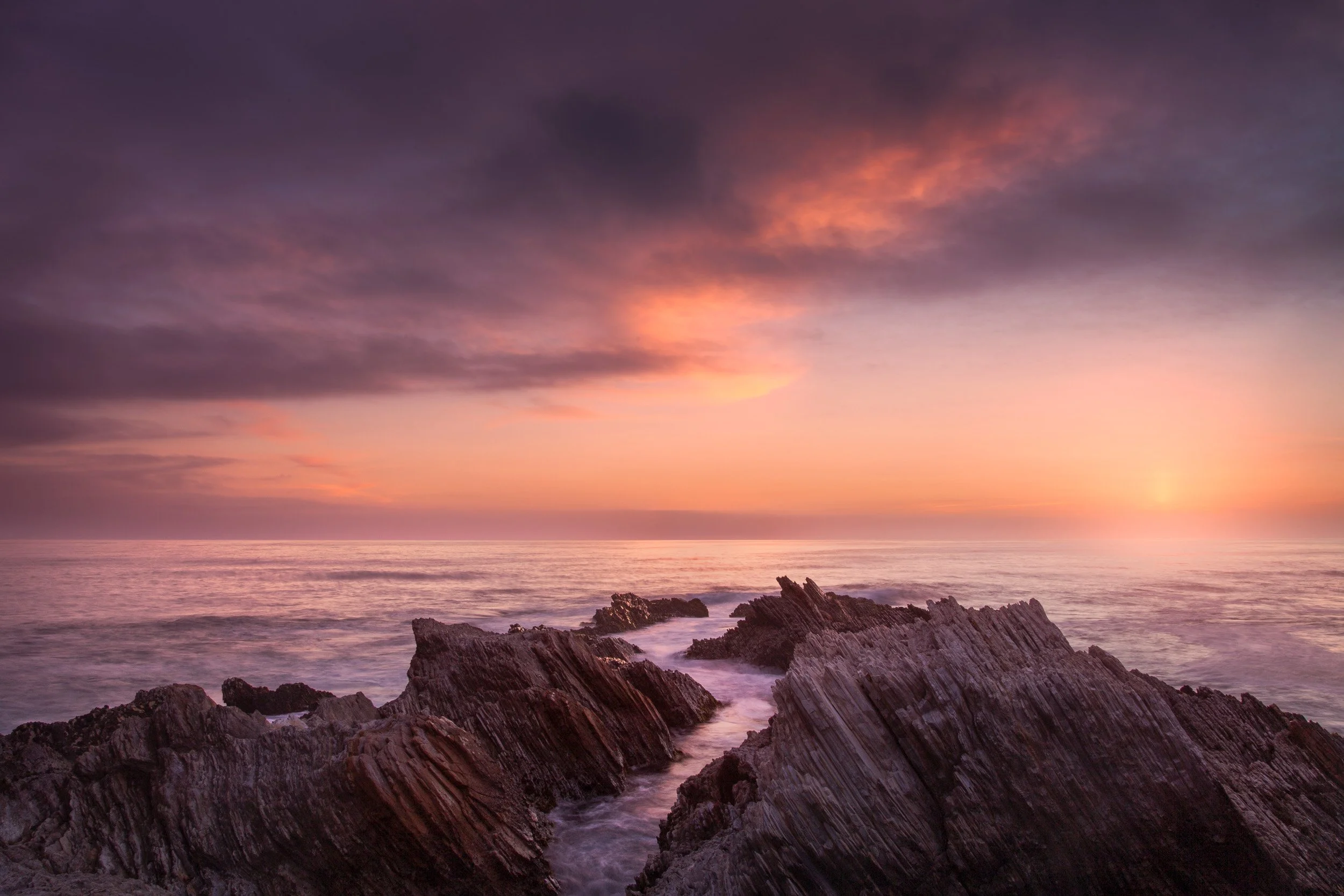 Tilted ridges of layered rock cut through smooth long-exposure surf beneath a pastel sunset, Montaña de Oro.