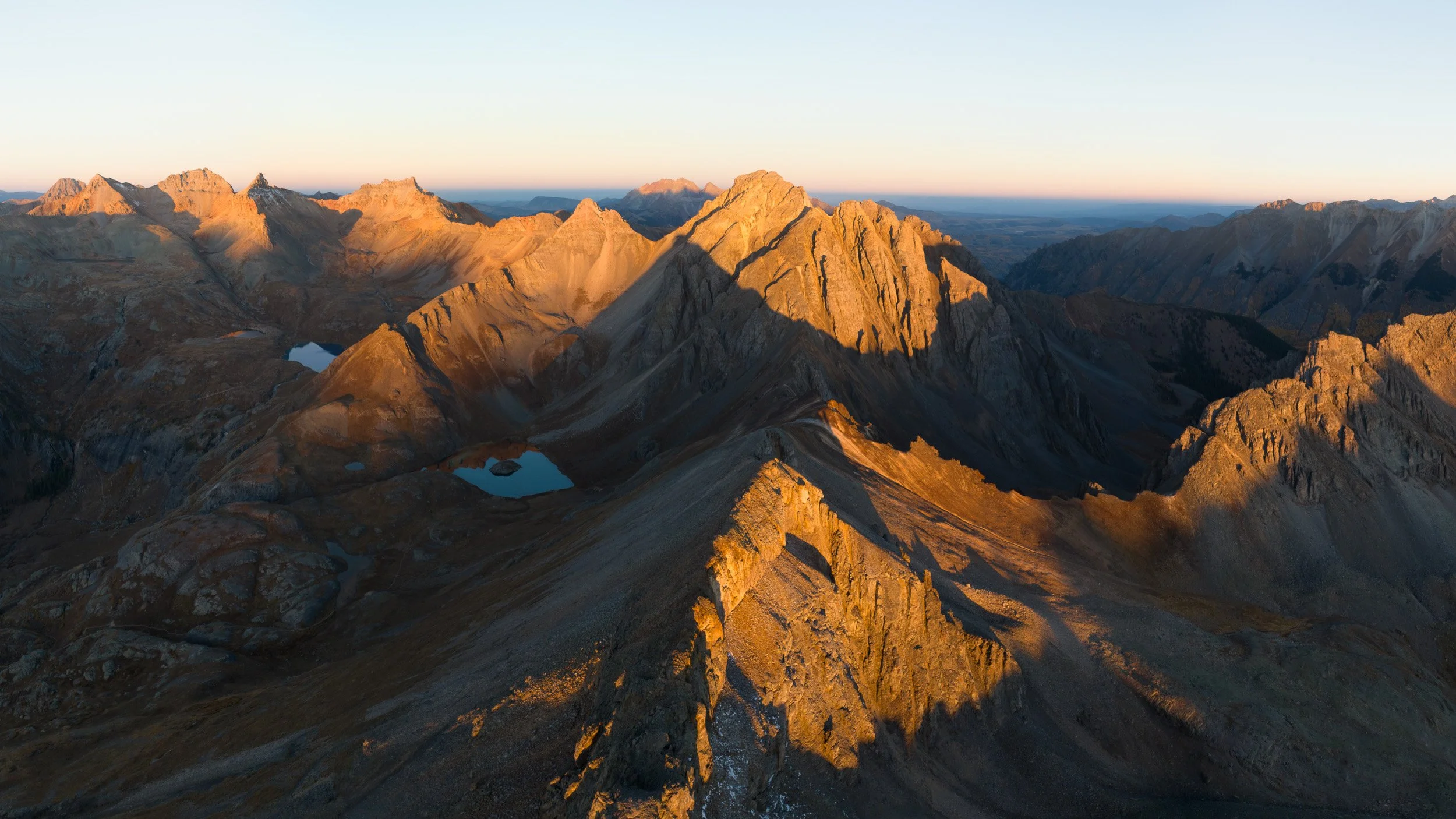 Aerial panorama of Colorado sunrise light raking across jagged San Juan peaks, with Island Lake and Ice Lake below.