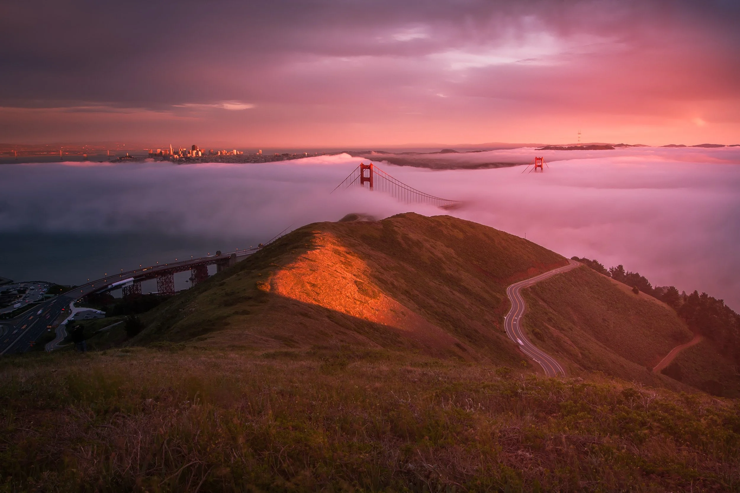 The towers of the Golden Gate Bridge rising above a sea of low fog at sunset, viewed from the Marin Headlands with San Francisco beyond.