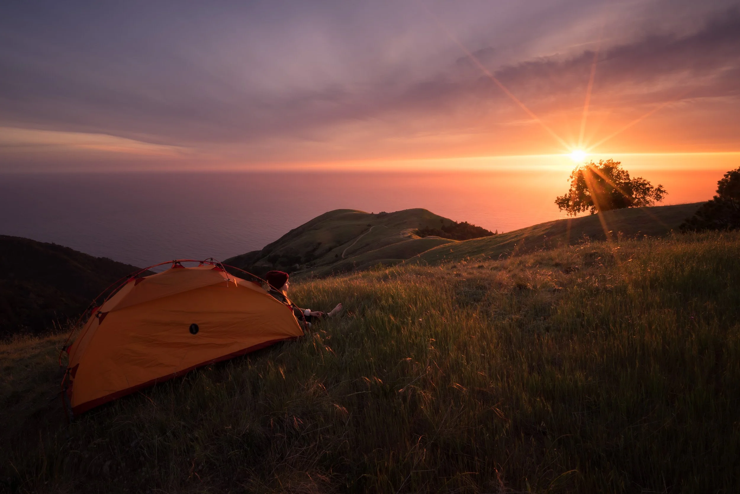 A camper beside an orange tent watches the sun set over the Pacific Ocean from a grassy ridge above the Big Sur coast, California.