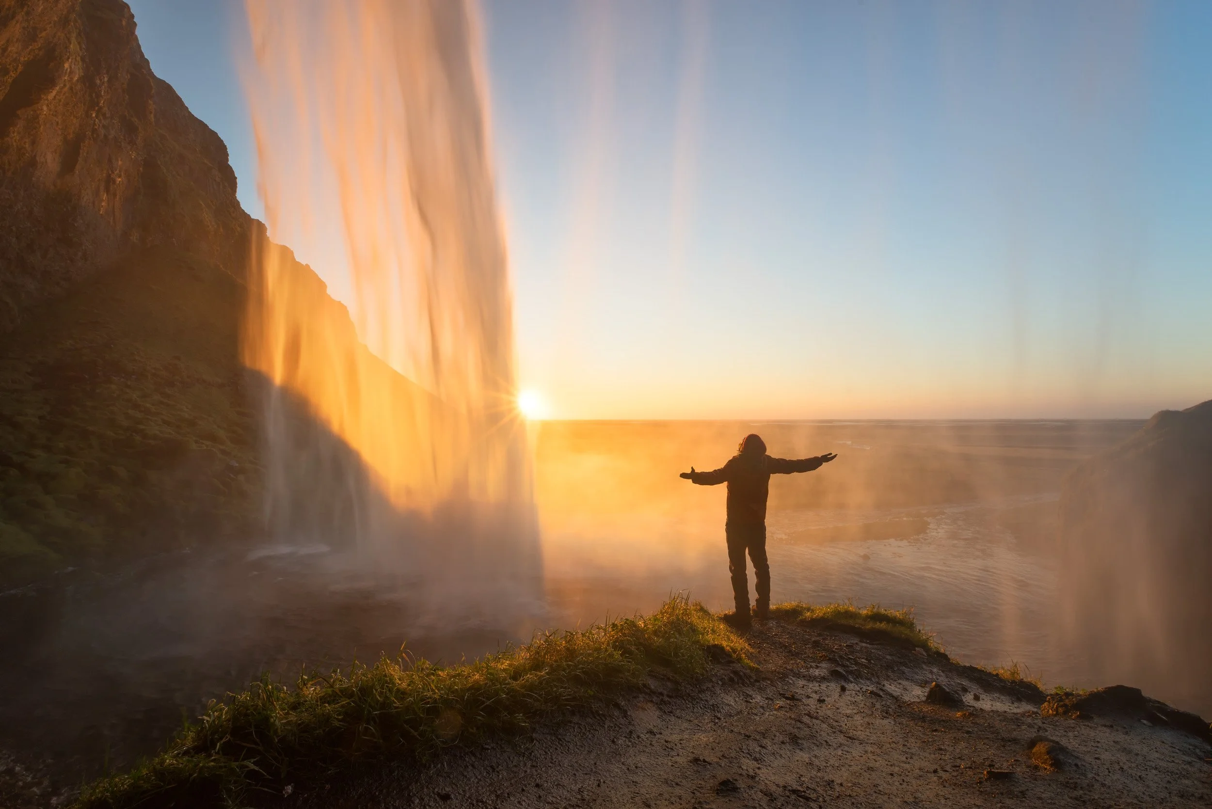 A person stands with arms outstretched behind Seljalandsfoss waterfall at sunset, golden light streaming through the falling water, South Iceland.