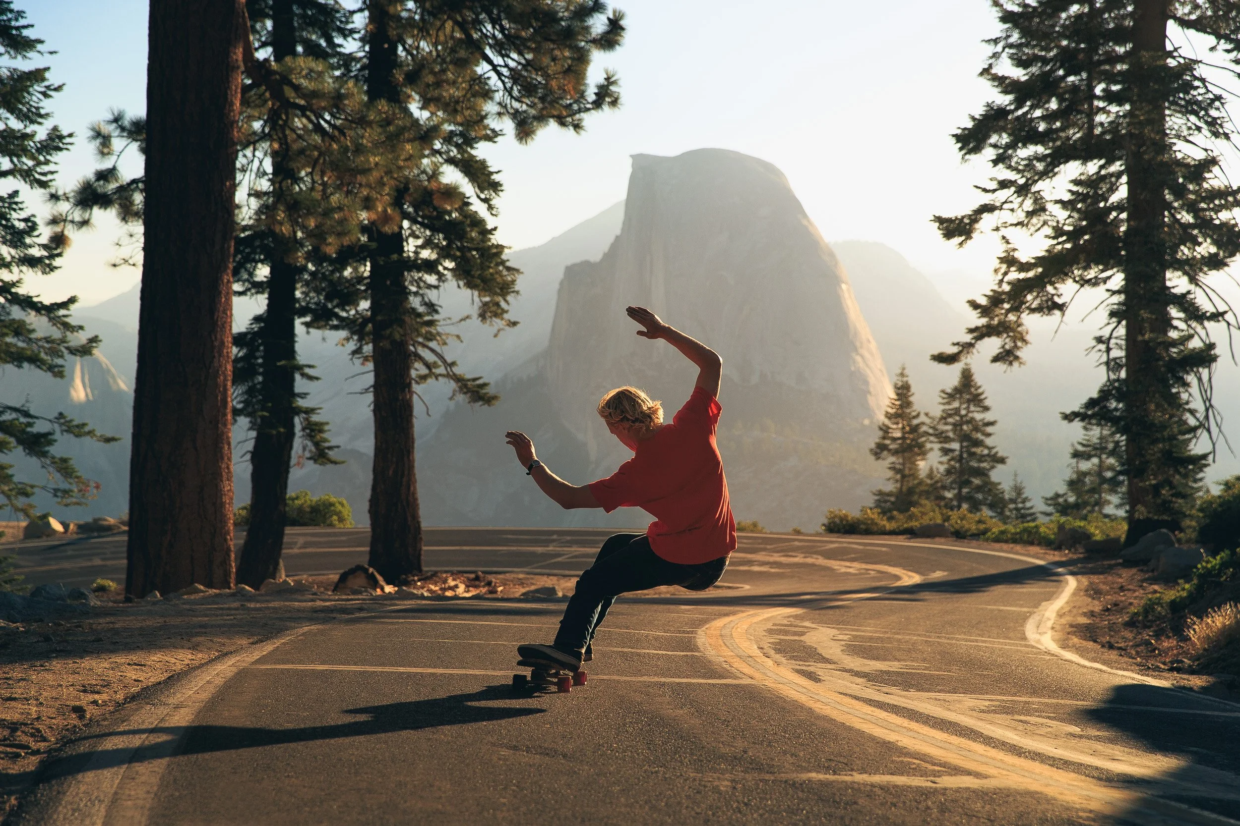 A skateboarder in a red shirt carves down a winding mountain road at Glacier Point with Half Dome rising in the background, Yosemite National Park.