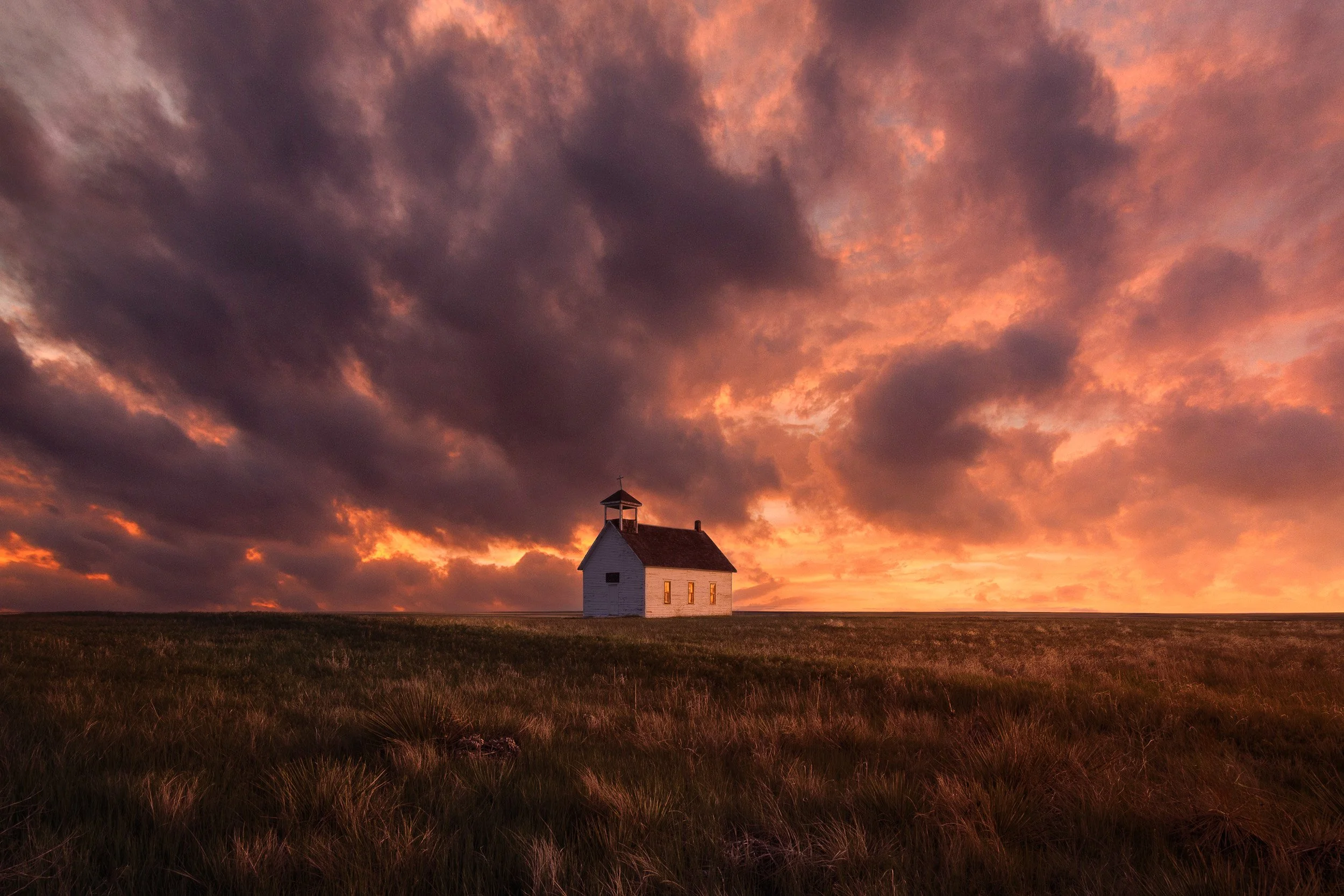A lone white wooden church with glowing windows under a dramatic pink and orange storm sky on the Colorado prairie at sunset.