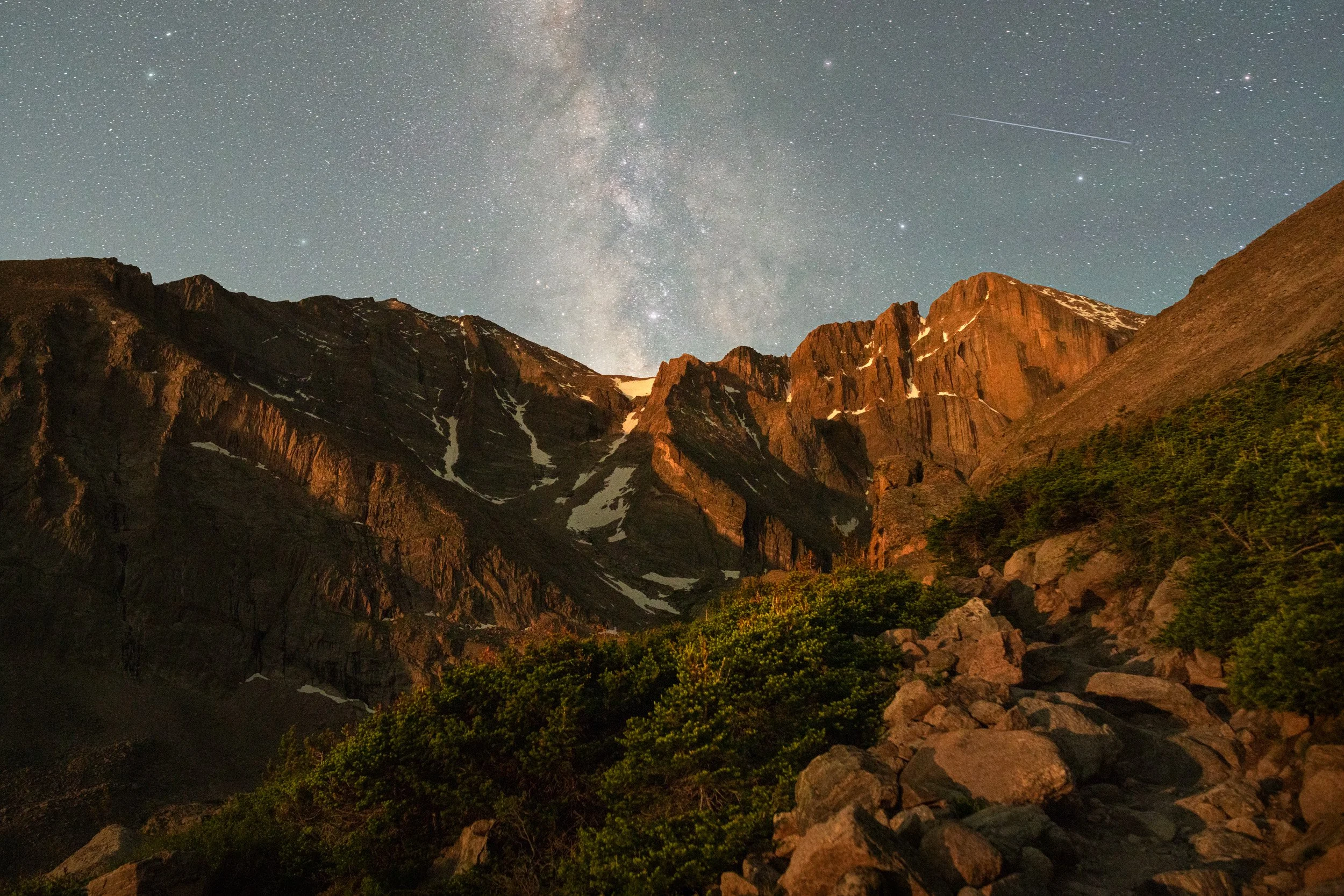 The Milky Way rises over the moonlit Diamond face of Longs Peak, with a meteor streaking above Chasm Lake trail, Rocky Mountain National Park, Colorado."