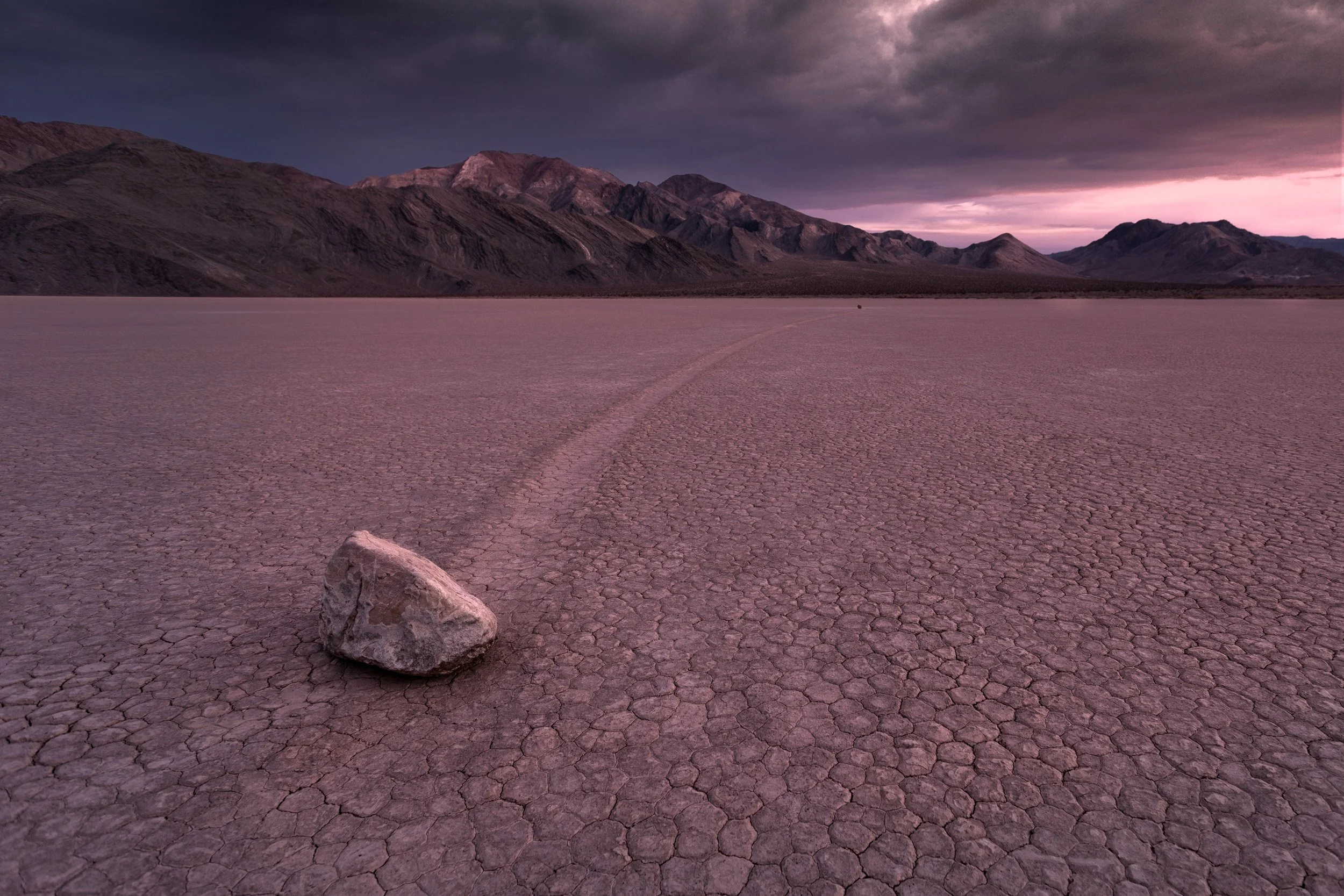 A sailing stone and its long track cross the cracked clay of the Racetrack Playa at dusk, Death Valley National Park.