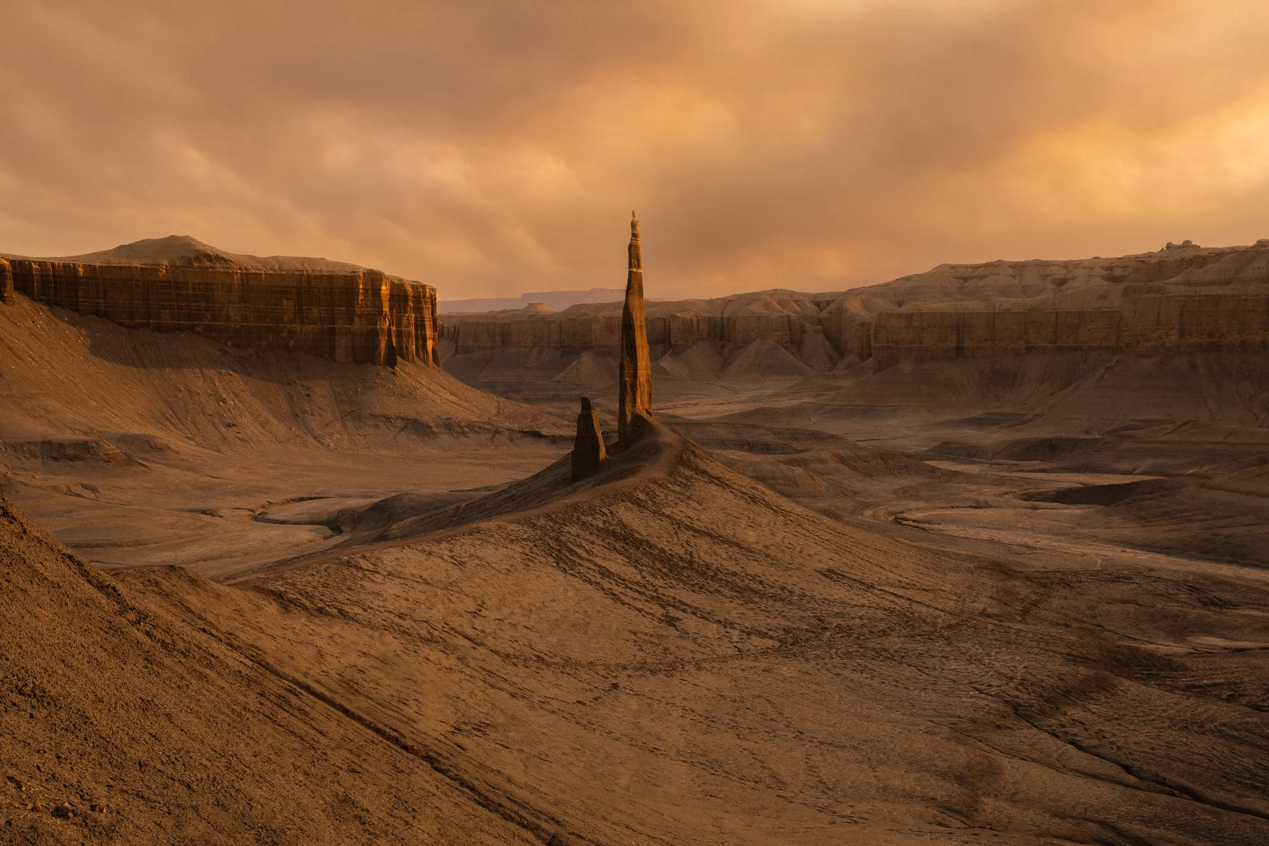 A slender rock spire rises from grey badlands below orange storm light near Hanksville, Utah.