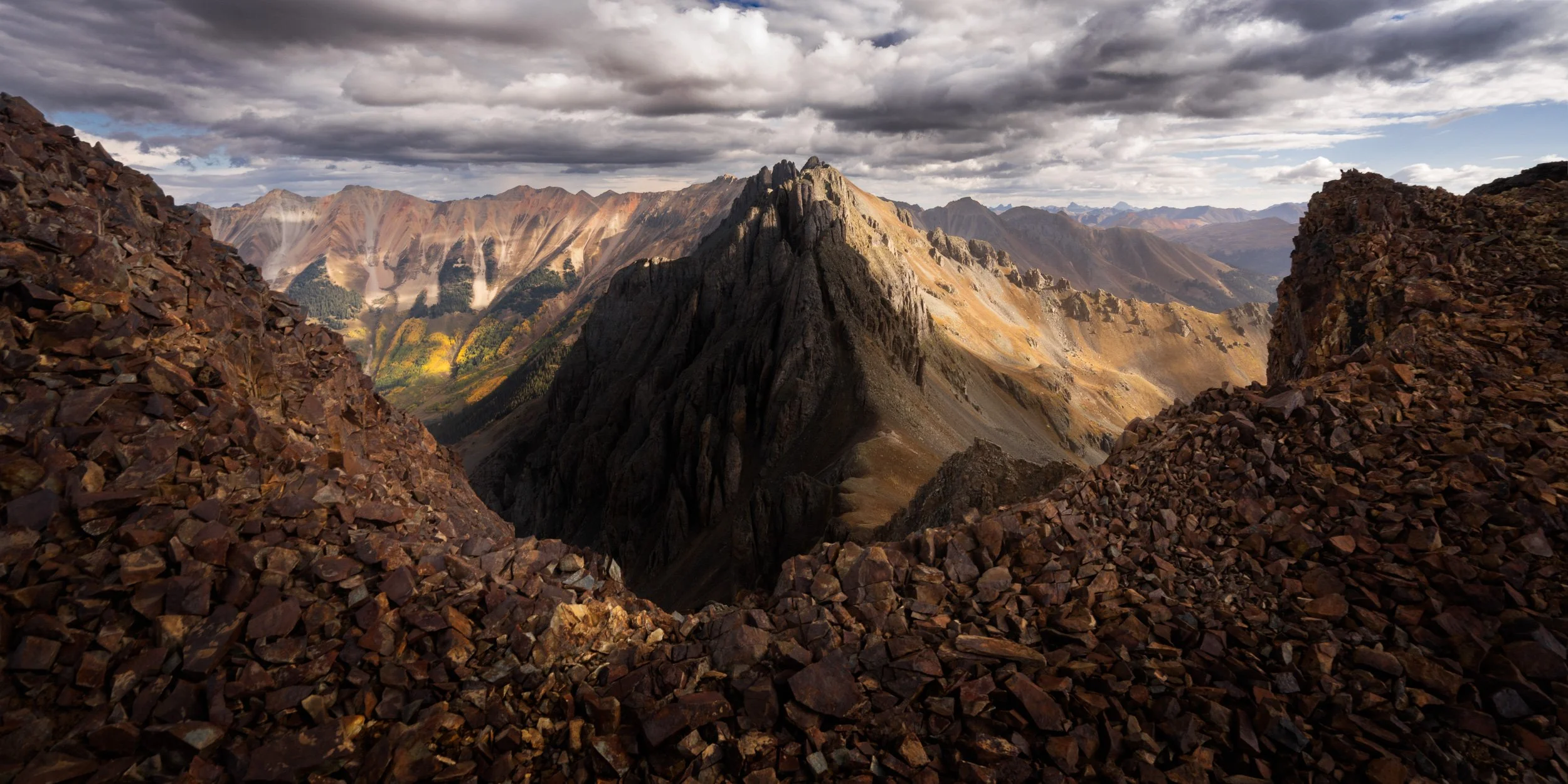 A panorama from a scree-covered summit ridge across jagged peaks and autumn valleys in the San Juan Mountains, Colorado.