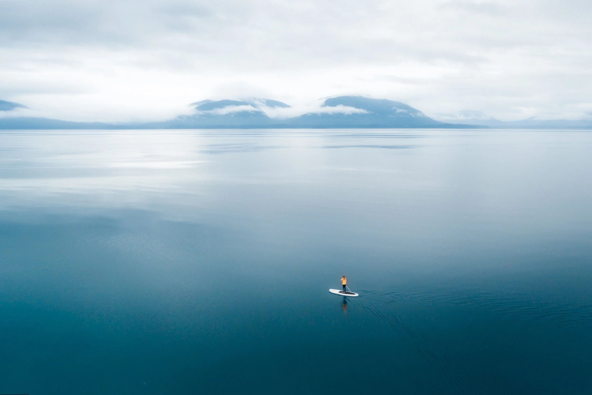 Aerial view of a lone paddleboarder in a yellow jacket crossing glassy blue water beneath fog-wrapped mountains, Alaska.