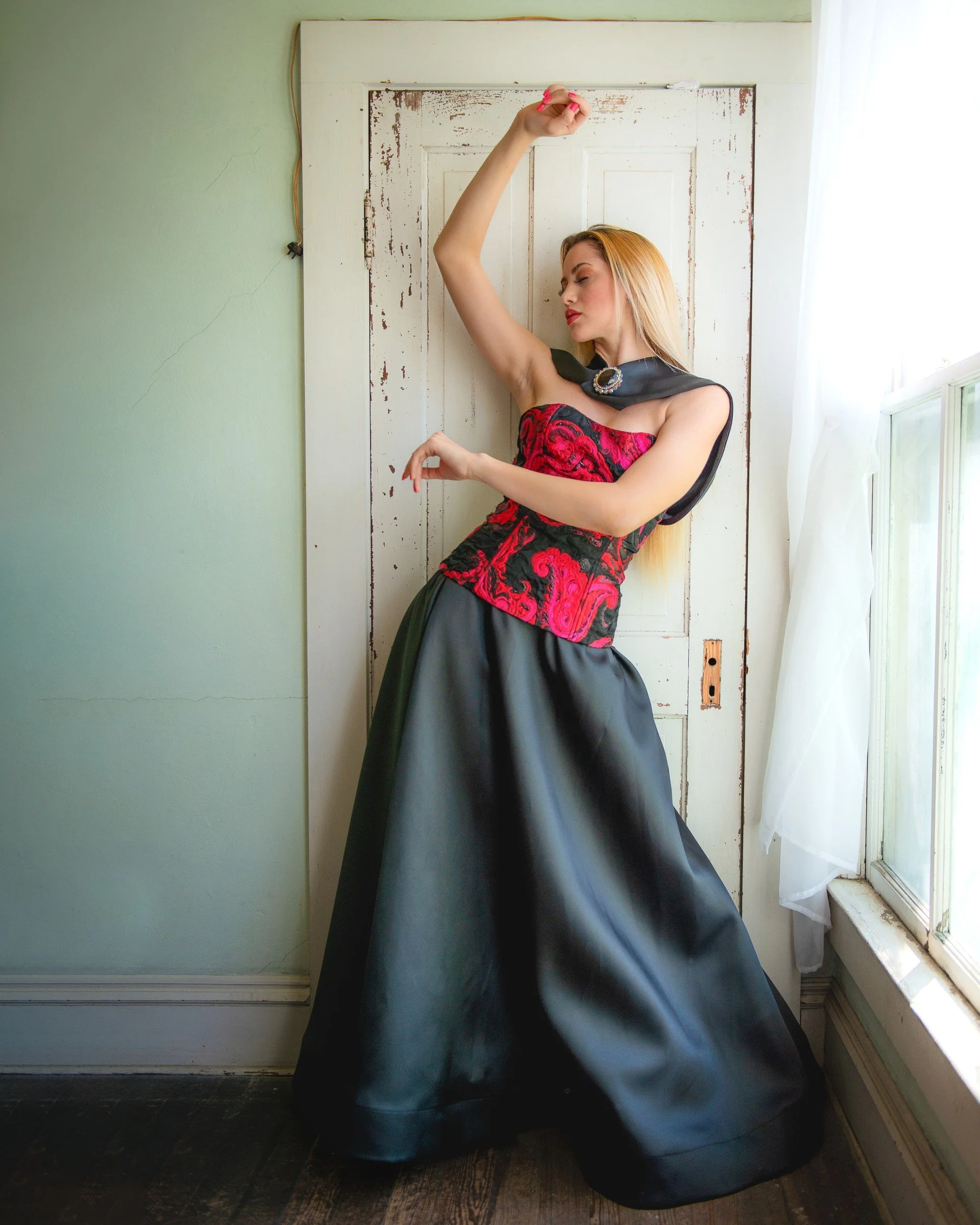 Model wearing a red and black gown from the Aurelia Batista collection beside a window. Photography by Zech Browning.