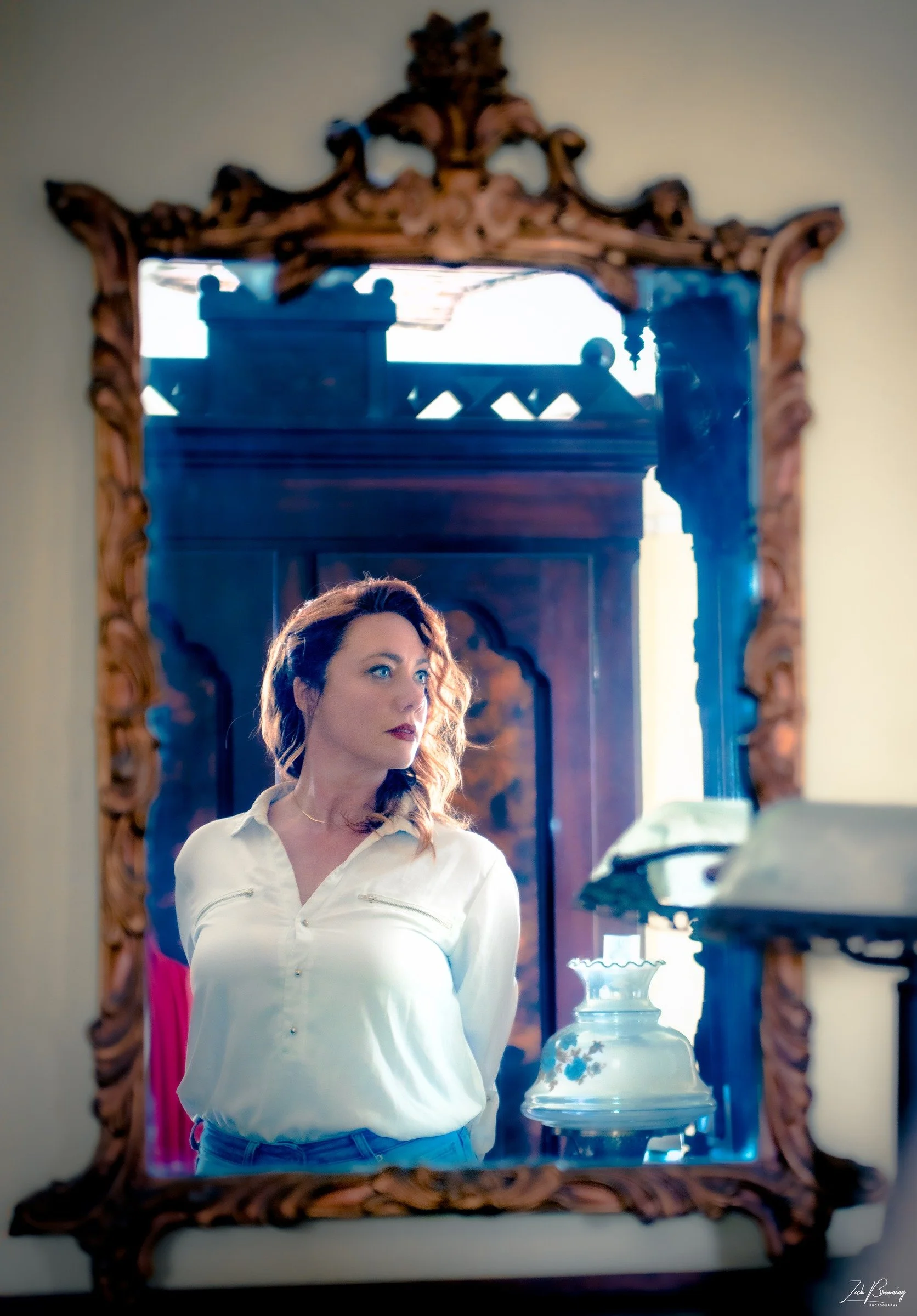 Dani in a white blouse, reflected in an ornate mirror with light catching her hair.