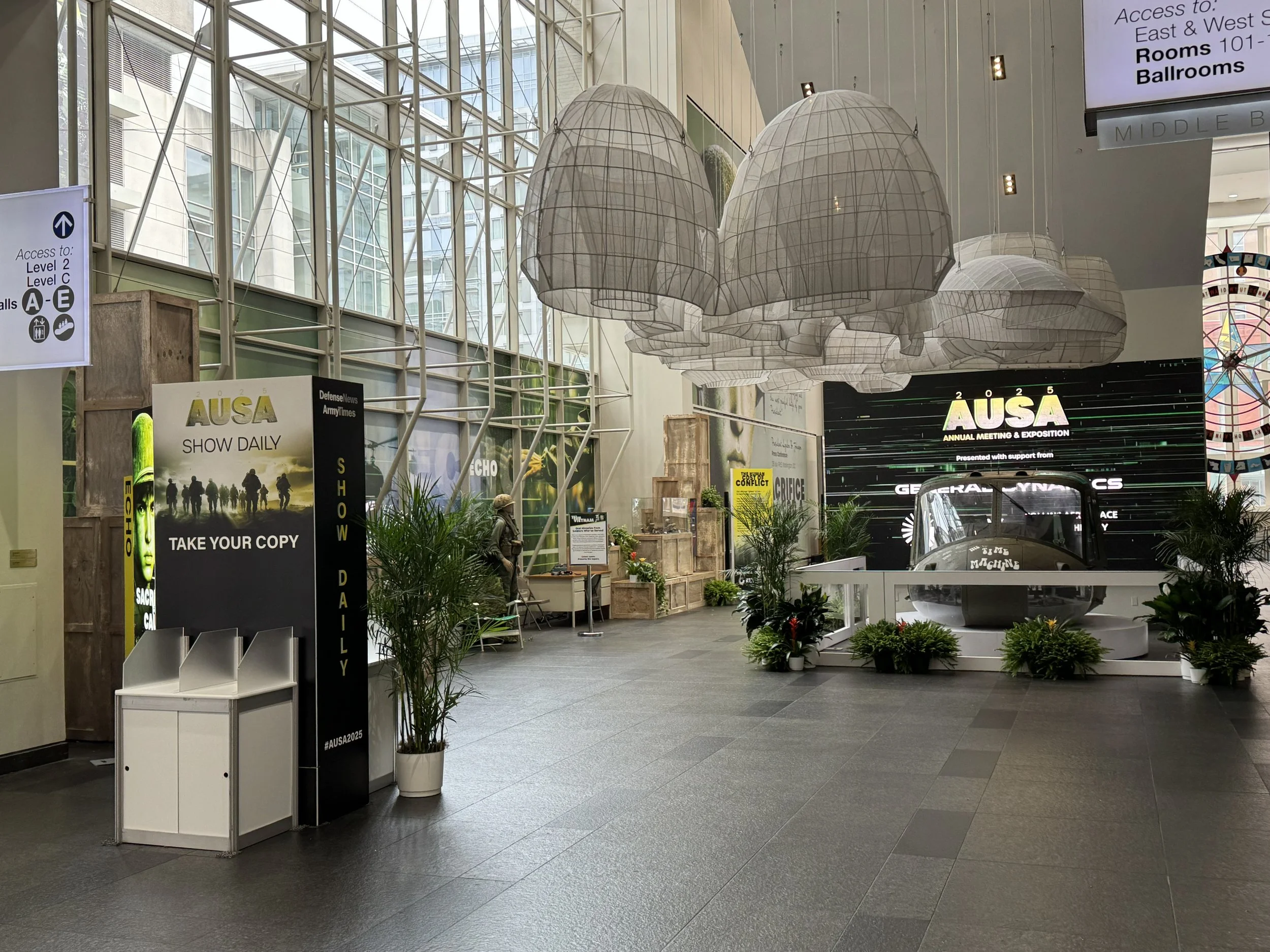 Interior of a large, modern convention center or airport terminal with high glass ceilings, decorative hanging lights, various signs for access and directions, plants, and a vintage vehicle on display.