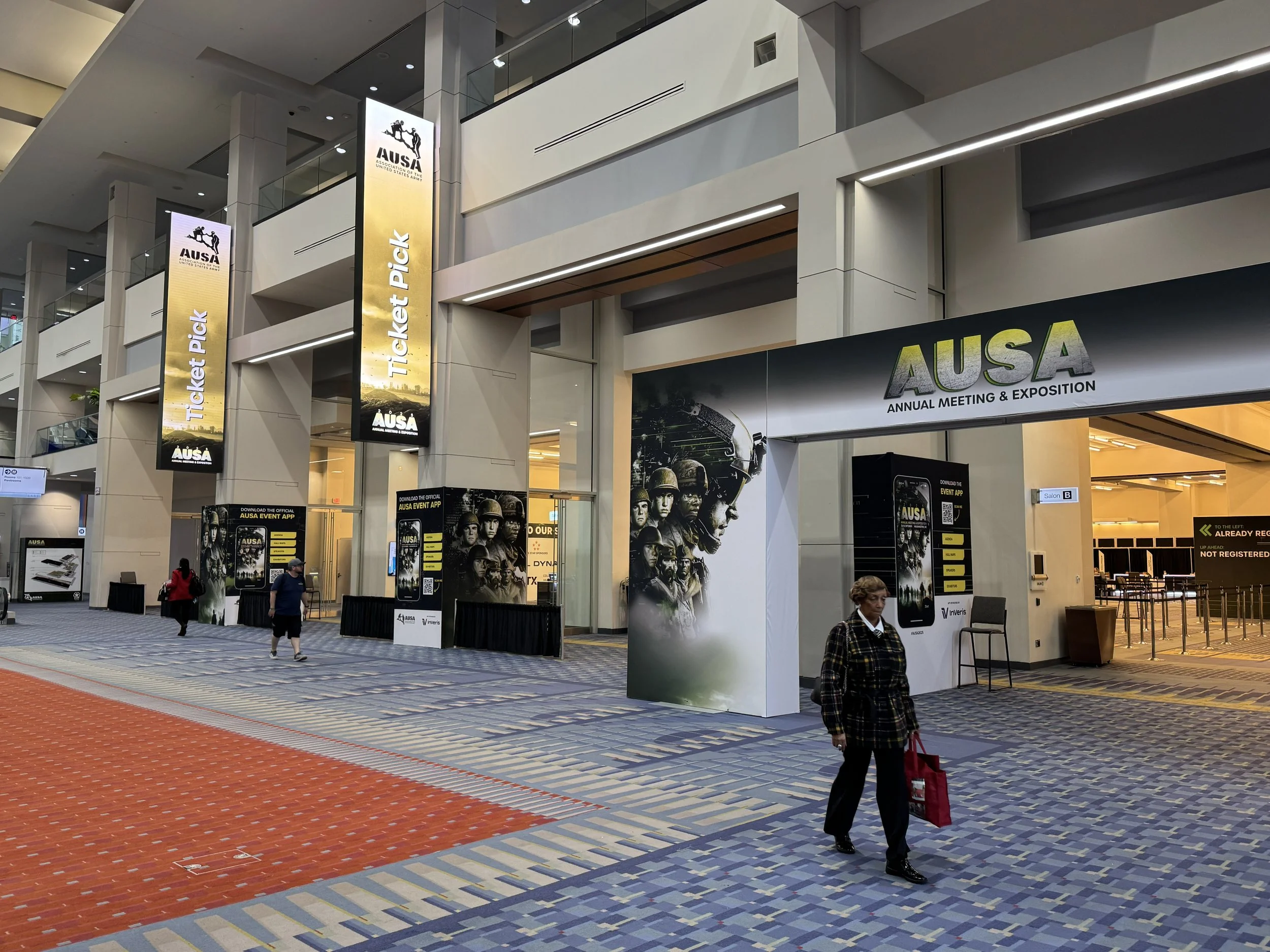 Interior of a large modern convention center with multiple floors, escalators, and banners advertising the AUSA annual meeting and exposition. Several people are walking around, some with bags, on a multi-colored carpet.
