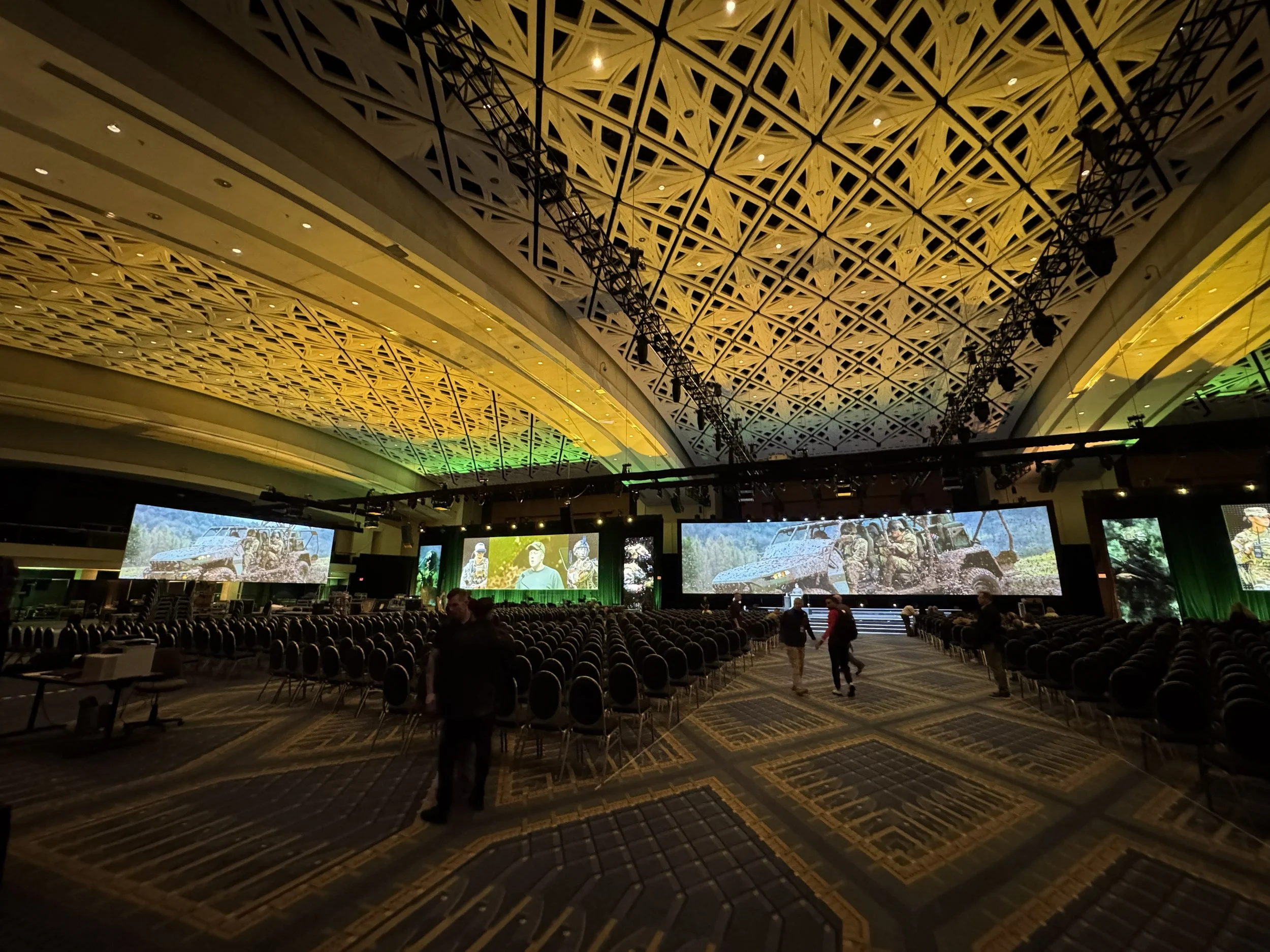 Large indoor space with a stage, multiple screens, rows of chairs, and decorative geometric ceiling panels illuminated with yellow and green lights. People are setting up or preparing for an event.