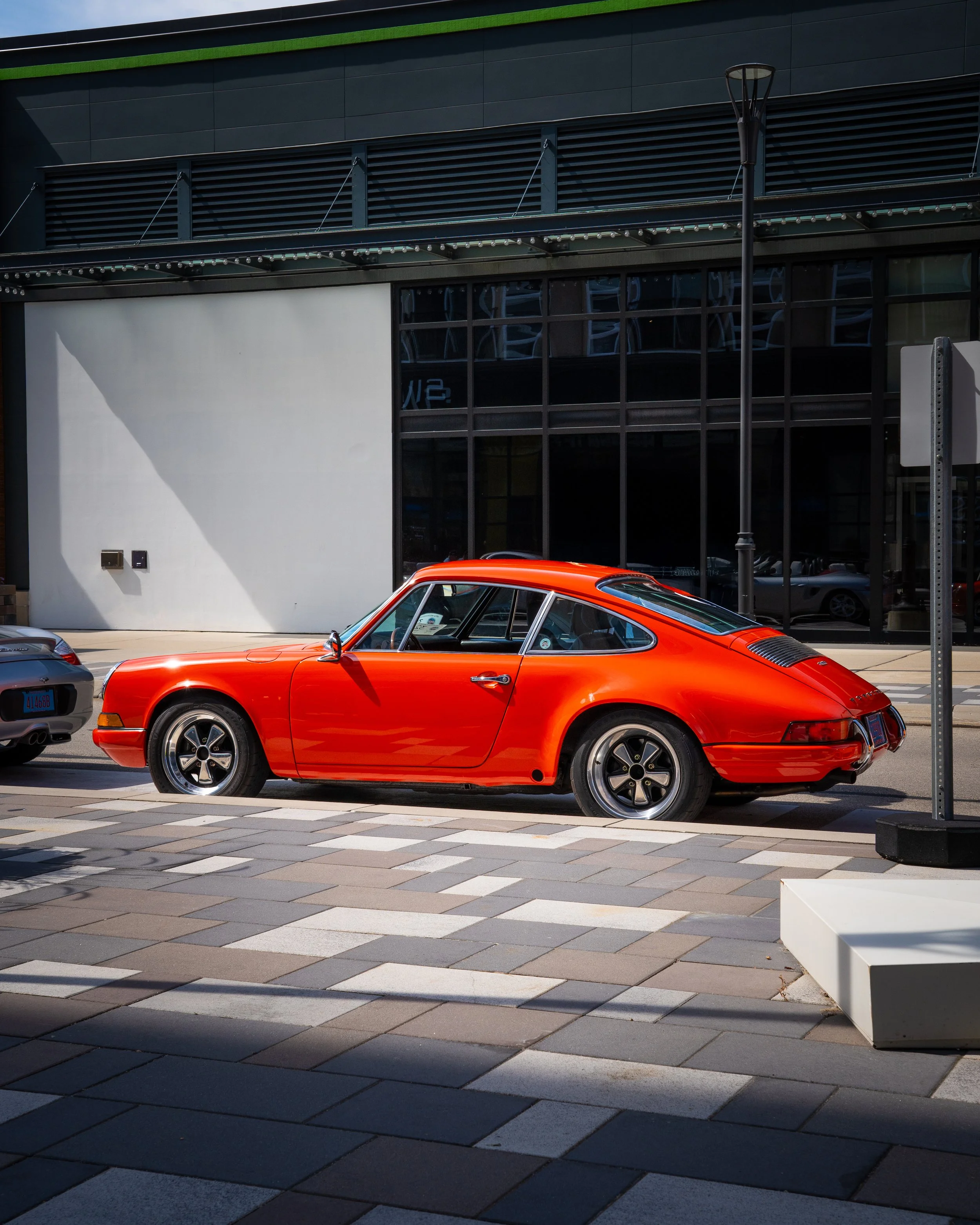 Red vintage Porsche sports car parked in front of a modern building with reflective windows and a sidewalk with tiled pavement.