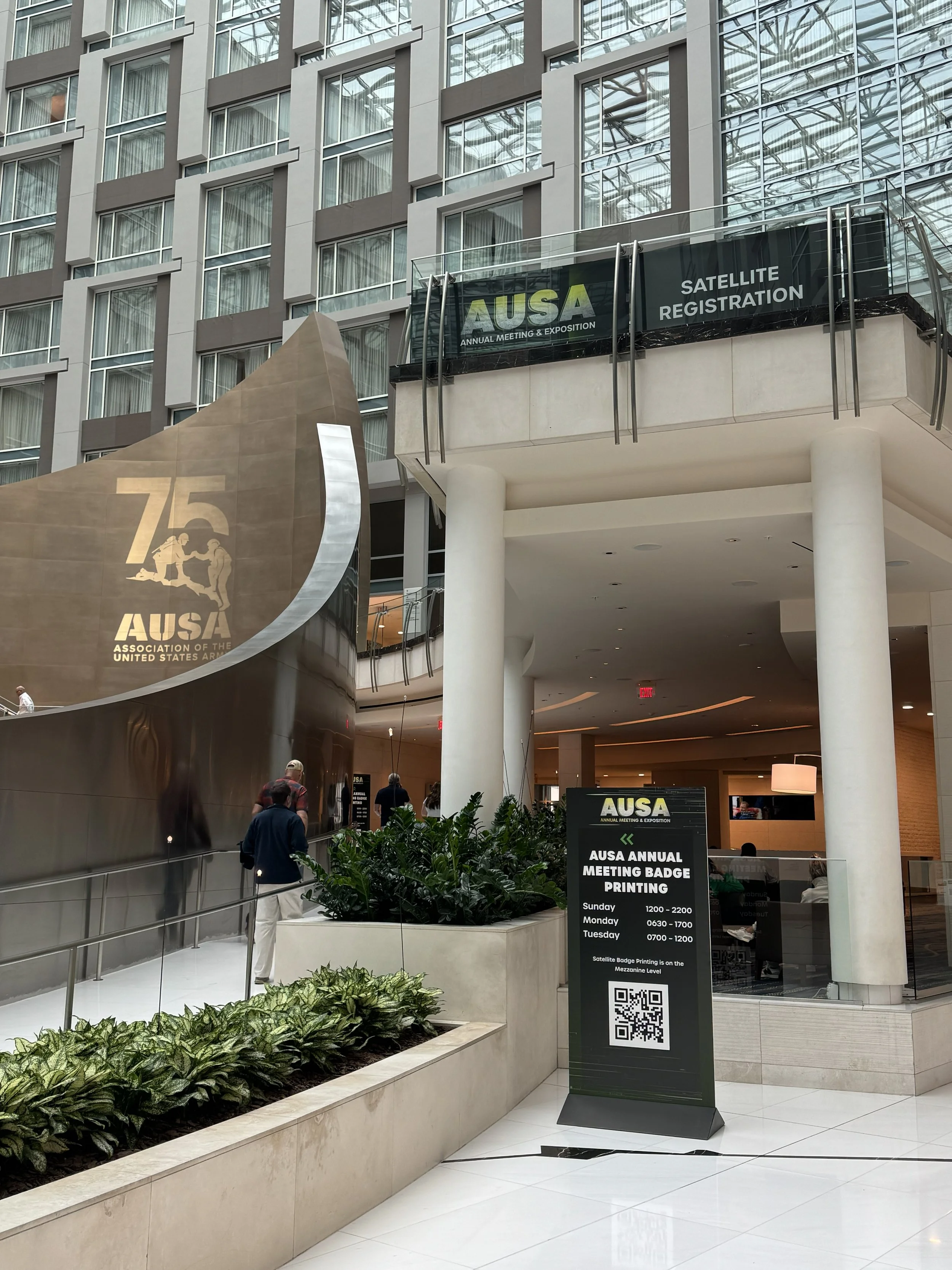 Interior of a modern building with a sign for the AUSA Annual Meeting & Exposition. People walk and gather near a sign that provides hours for badge printing, with plants and greenery in the foreground and a metal and glass architectural design in th