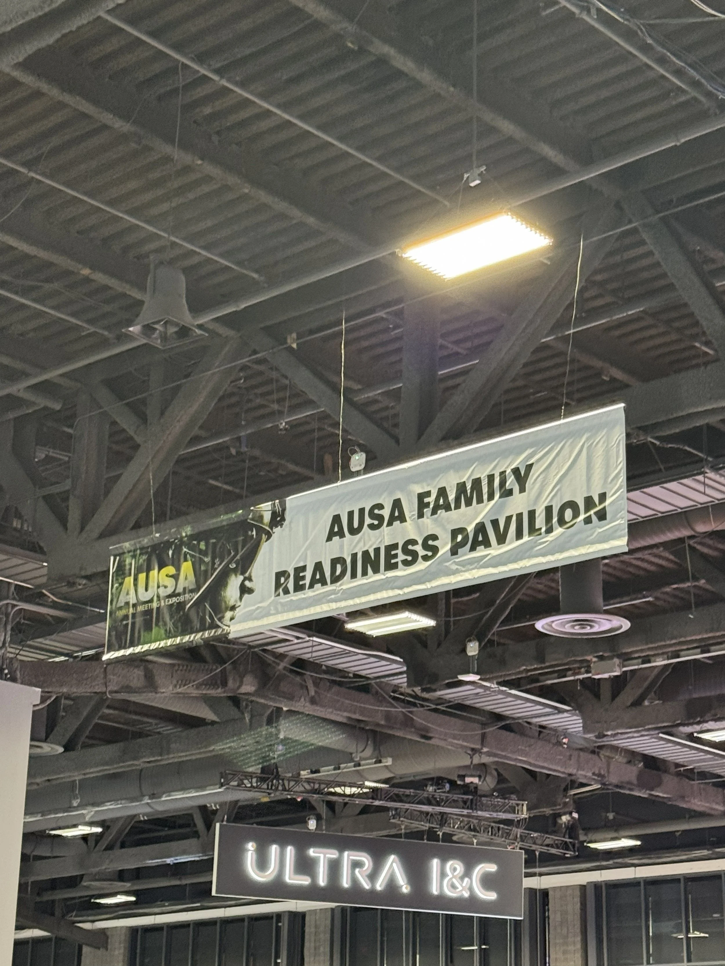 Interior of a convention center with hanging banners and signage. One banner reads 'AUSA FAMILY READINESS PAVILION,' and there is a neon sign below that says 'ULTRA 1&C.'