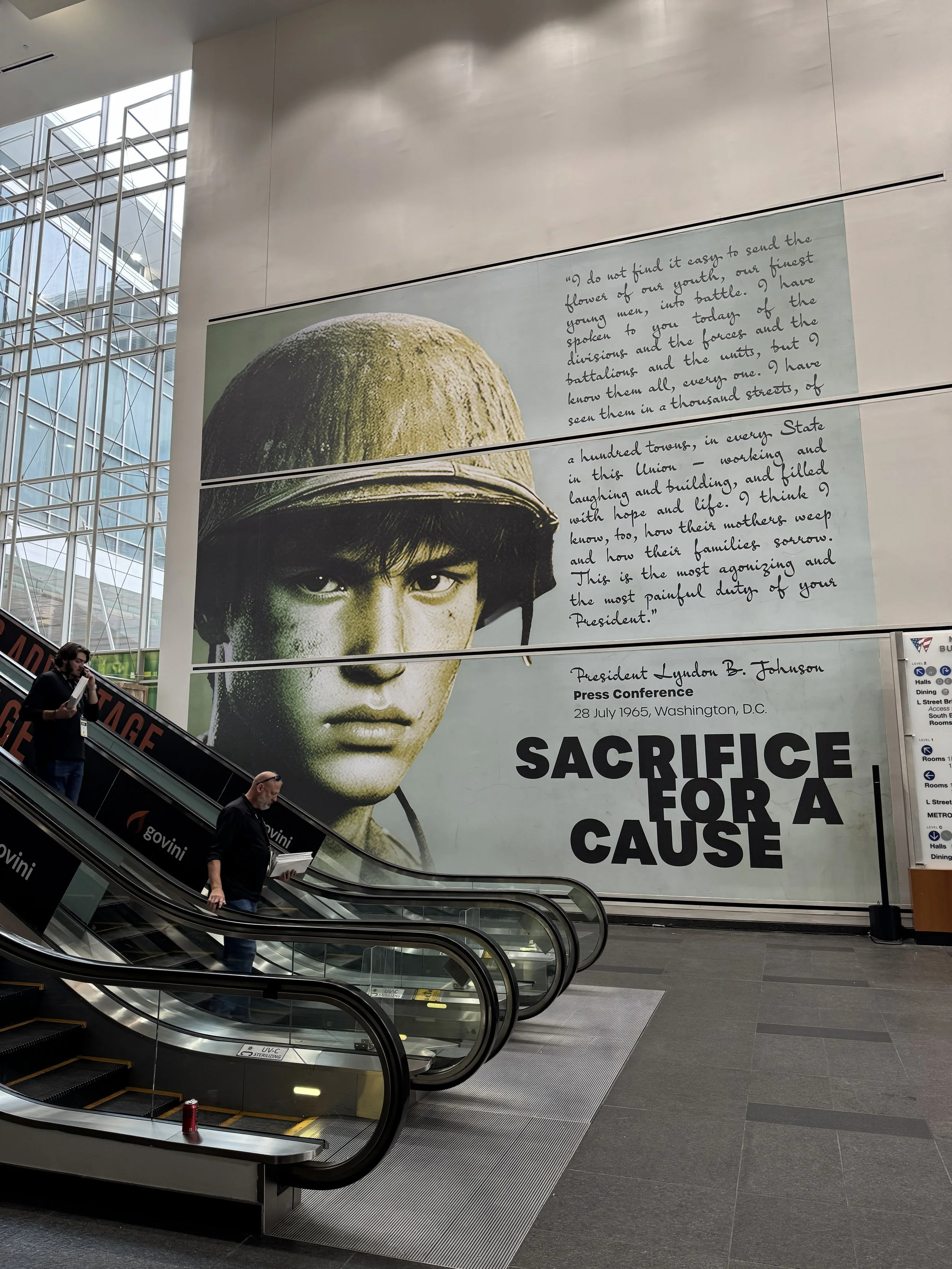 A large poster inside a modern building with a portrait of a soldier wearing a helmet and a quote written in cursive. The quote discusses sacrifice and duty. Two people are seen on an escalator in the foreground.