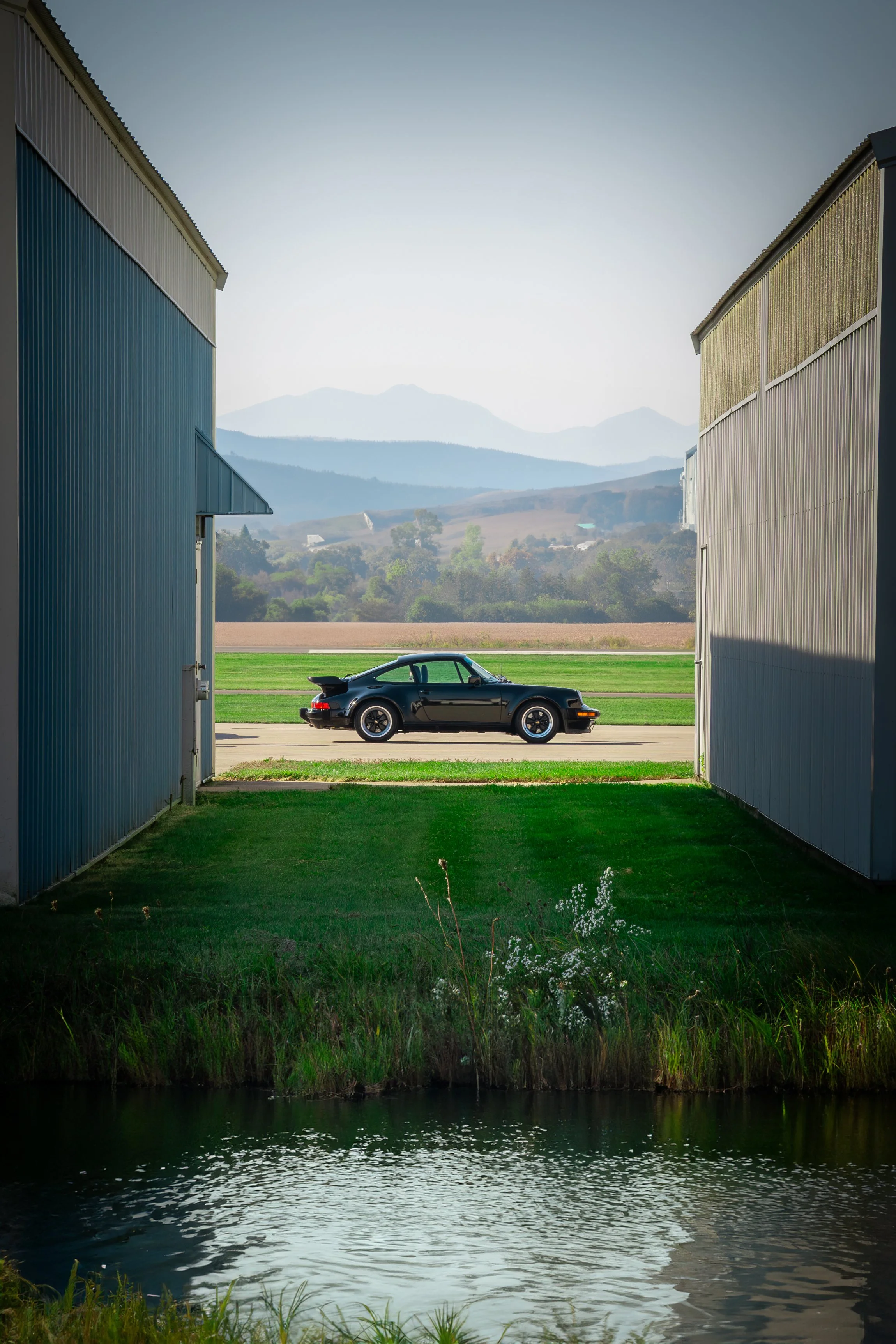 A vintage black Porsche sports car parked outside between two large blue and gray metal industrial buildings with a scenic mountain backdrop and a pond in the foreground.