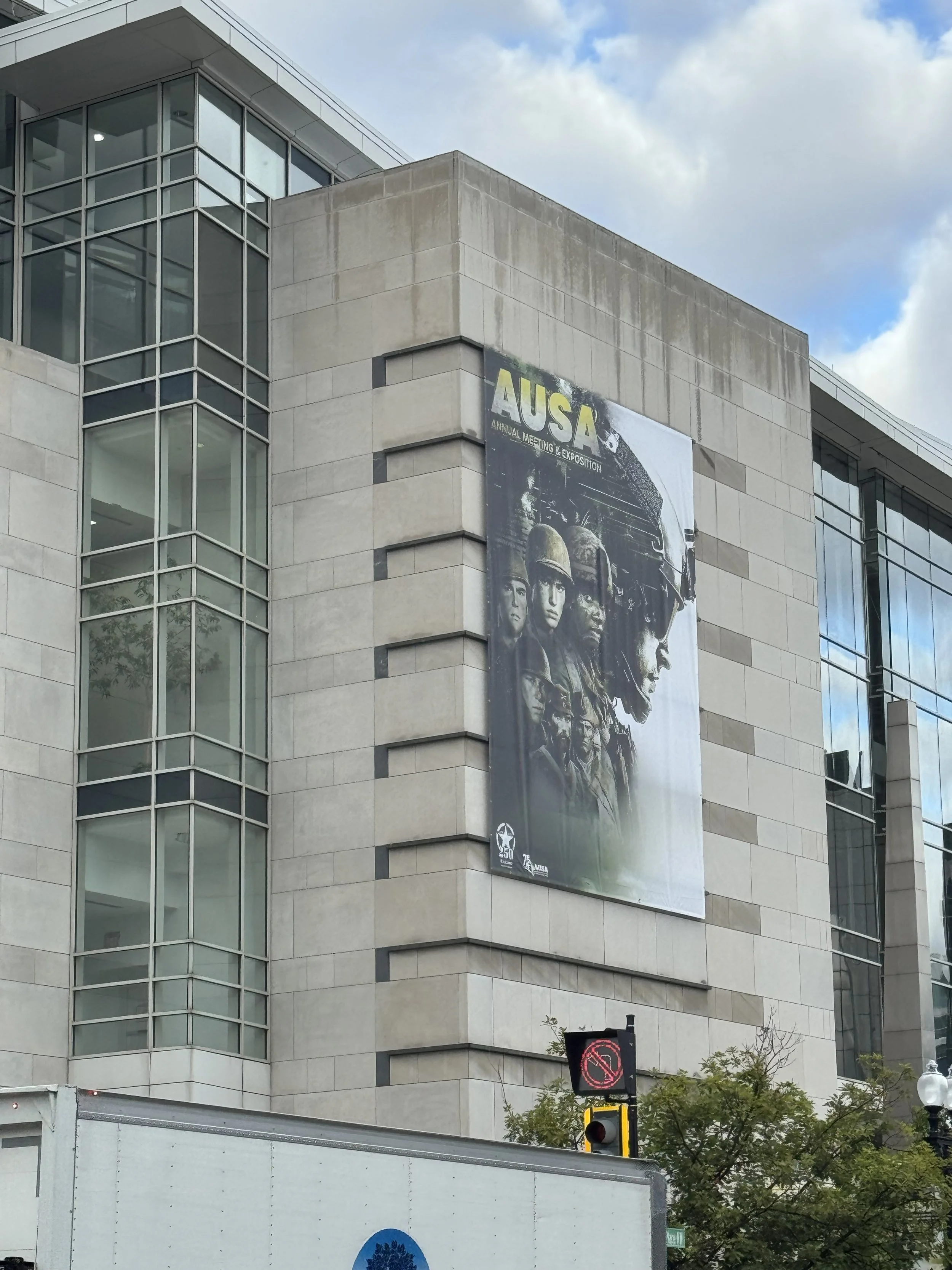 A large building with a modern glass and concrete exterior. A prominent banner displays the logo and soldiers for the AUSA (Army Officers Association) Annual Meeting & Exposition. The sky is partly cloudy.