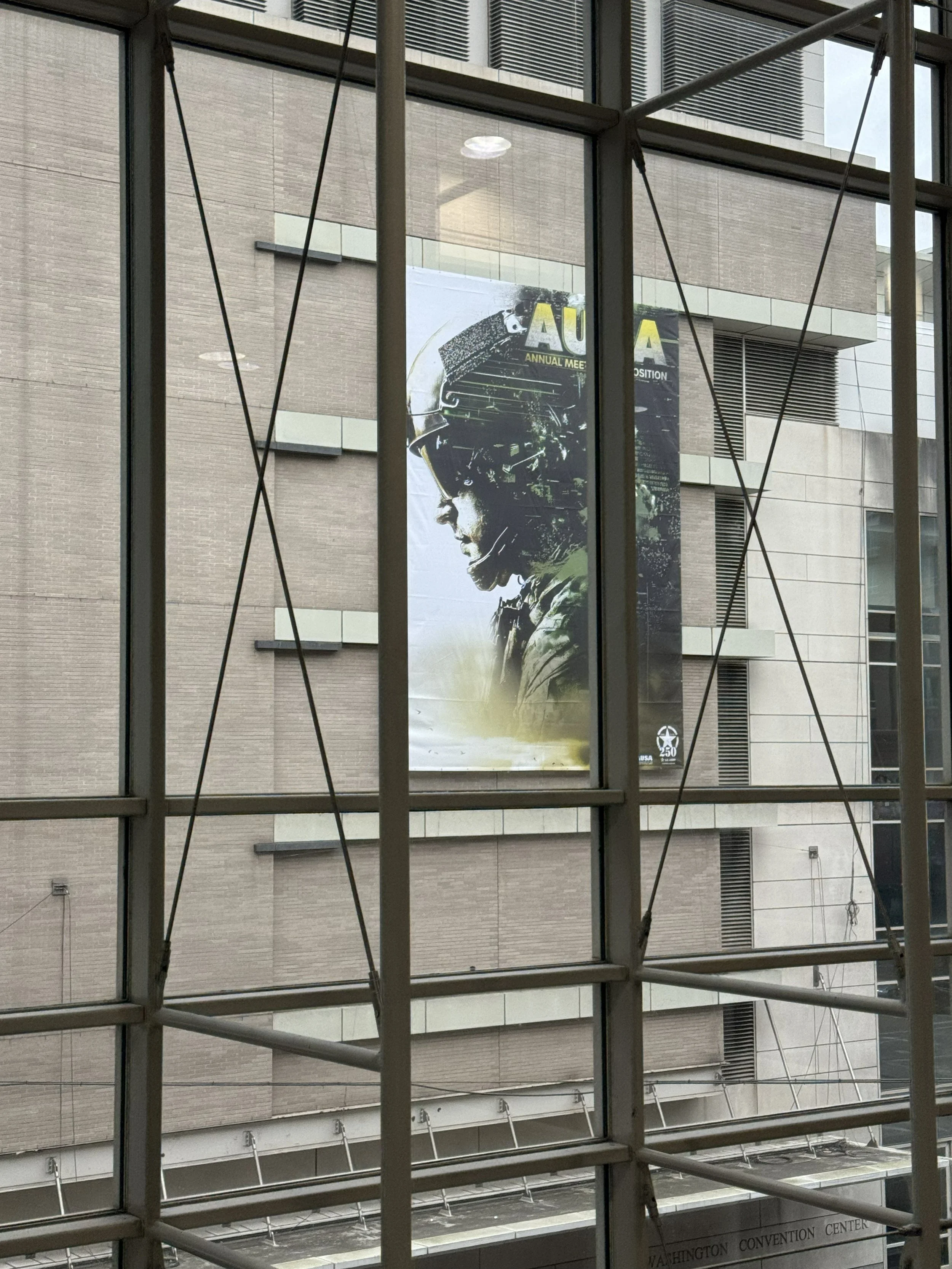 Photo taken from inside a building, looking through a large grid-style window at a poster on a building across the street. The poster features a side profile image of a person wearing a helmet. The background of the poster has text that says 'AUSA An