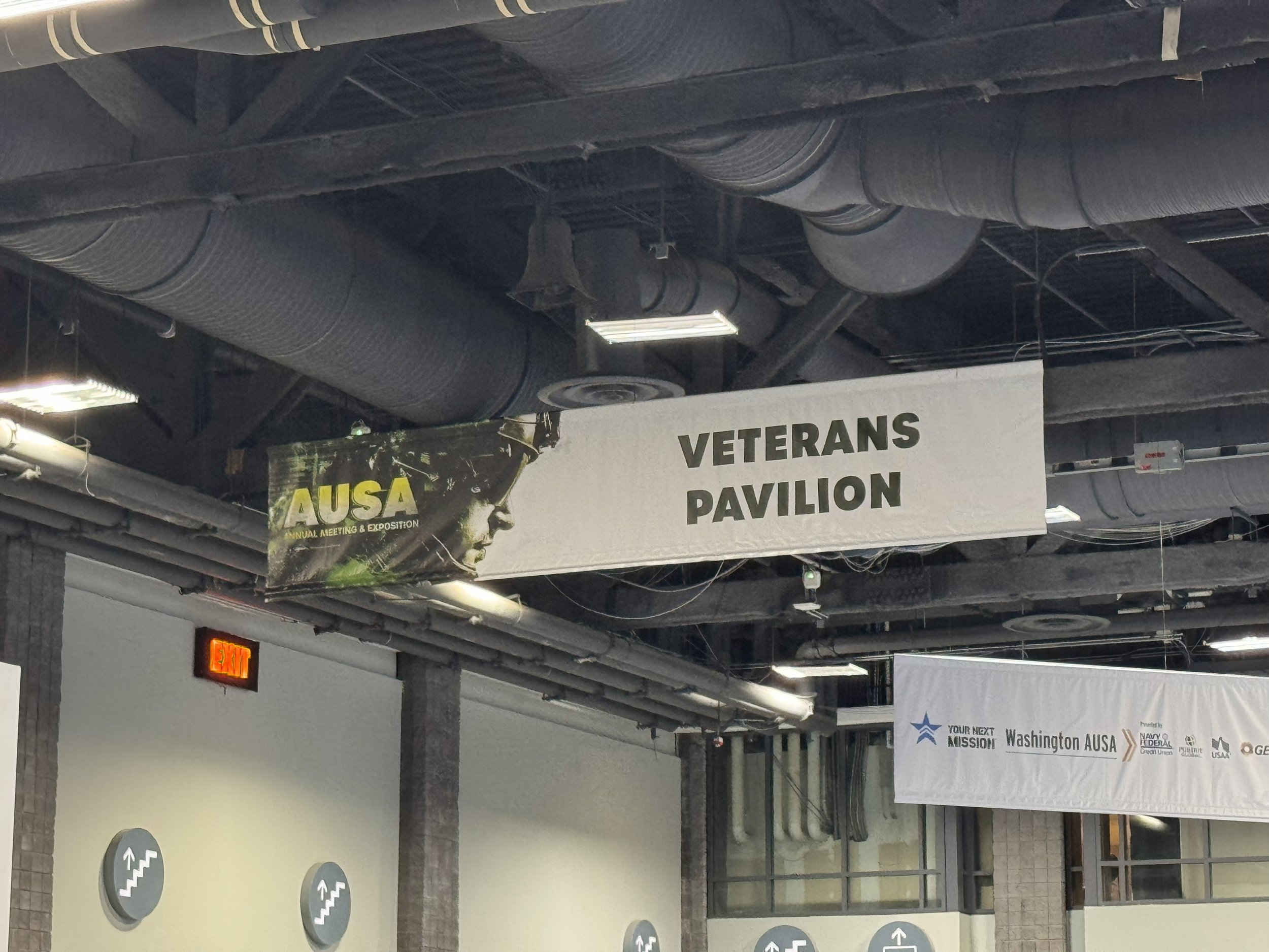 Indoor area with pipes on the ceiling, signage indicating 'Veterans Pavilion' and posters for a conference or event related to Washington AUSA, with directional symbols for stairs and information signs, and an illuminated 'Exit' sign.