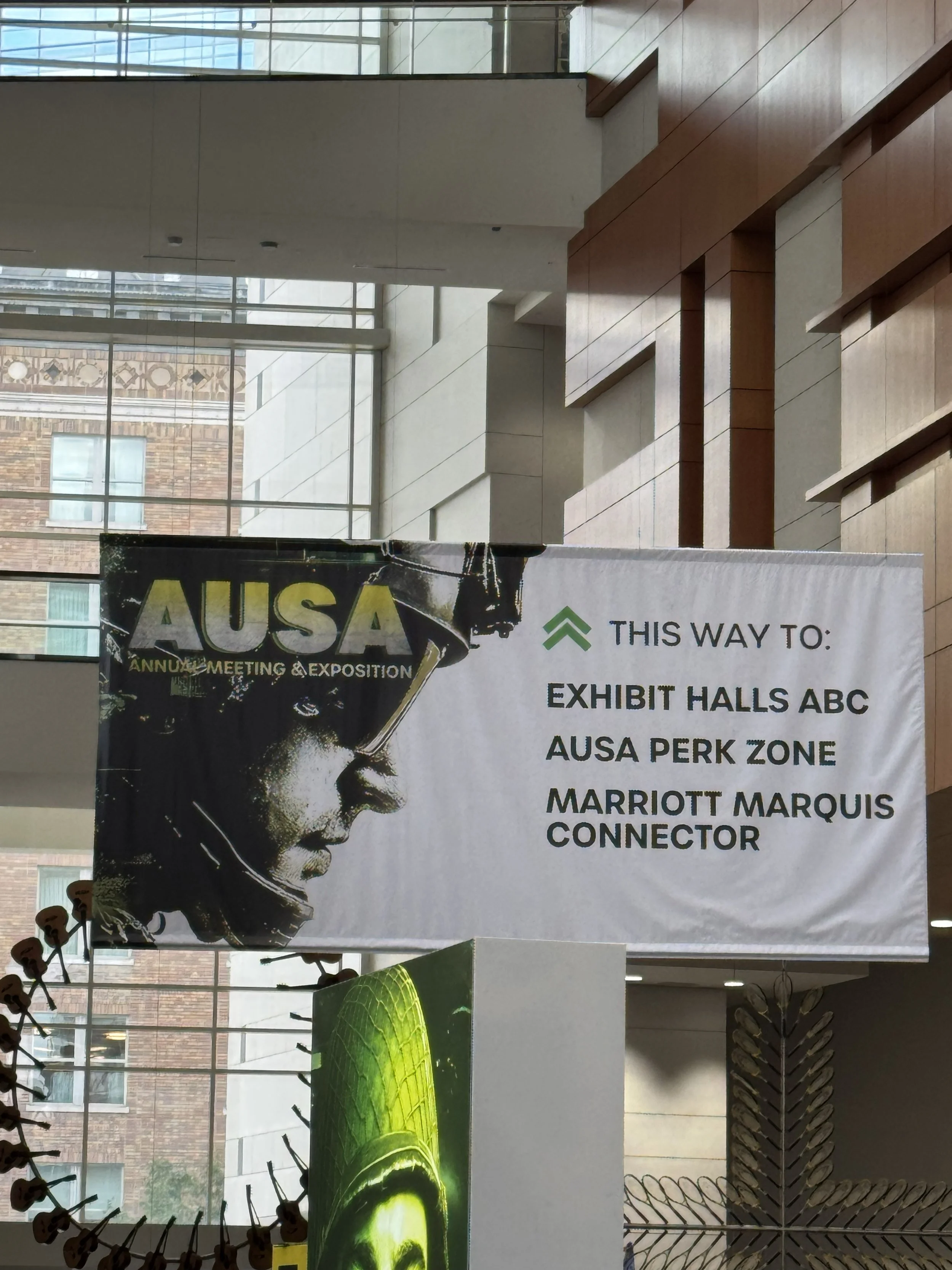 Indoor view of a large banner at a conference or exhibition with the text: 'THIS WAY TO: EXHIBIT HALL 10, AUSA PERK ZONE, MARRIOTT MARQUIS'. The banner features a portrait of a woman in profile with eye makeup, along with a green peace dove logo.