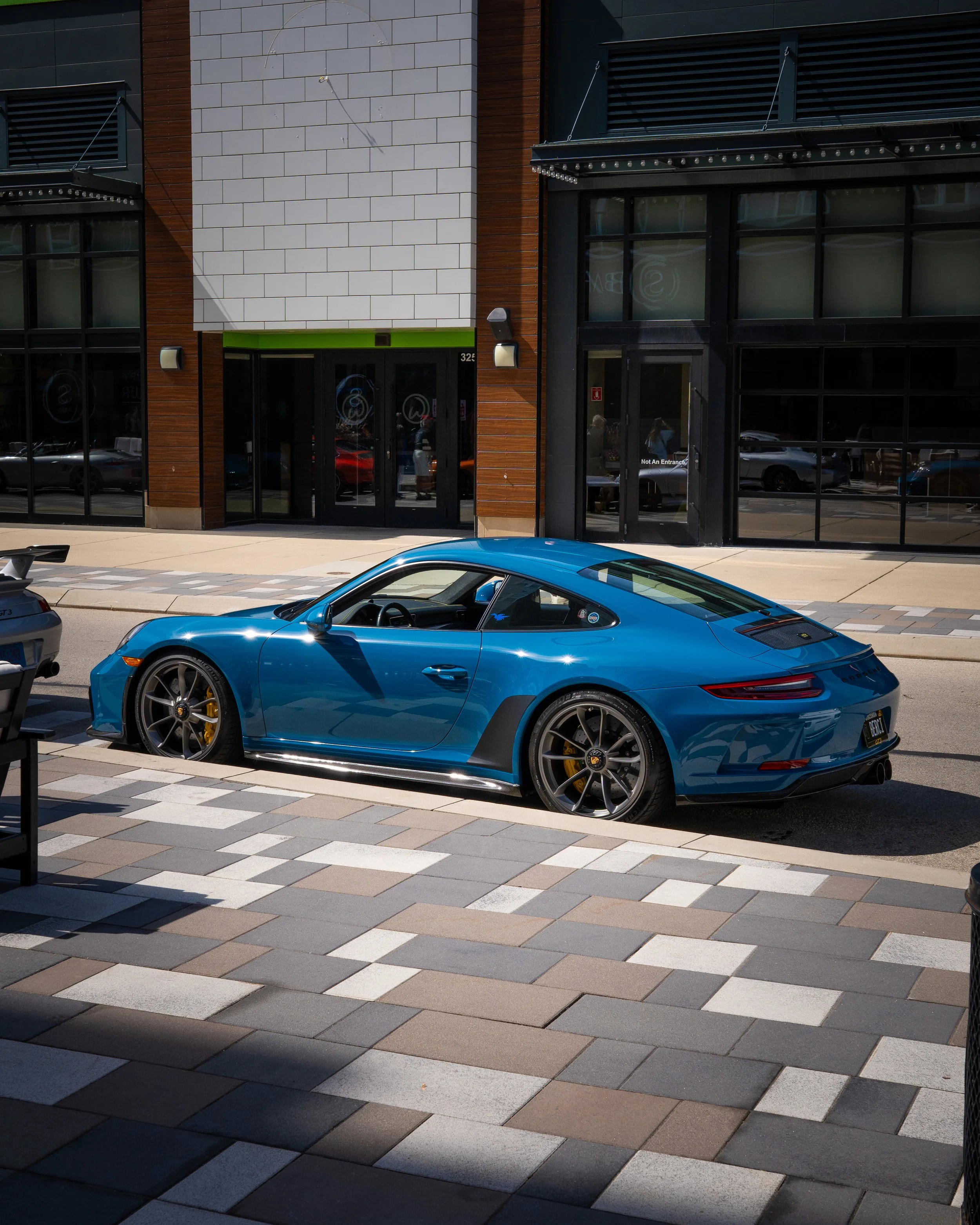 A modern building with a mix of brown and black exterior walls, large glass windows, and a sliding glass door. In front, there's a parking lot with a bright blue sports car parked in the foreground, and other cars visible through the windows and refl