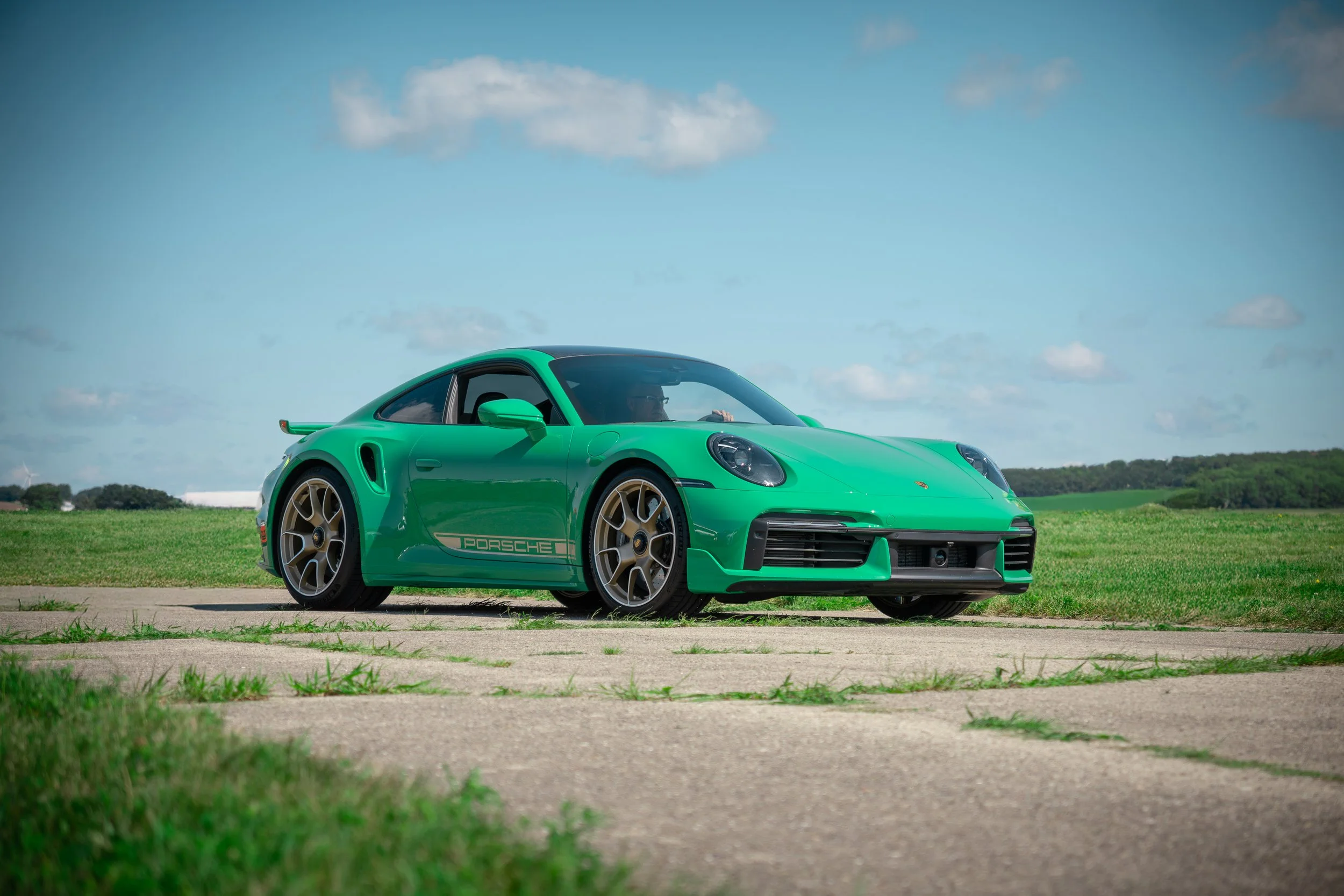 A bright green Porsche sports car parked on a concrete road in a grassy field under a cloudy blue sky.