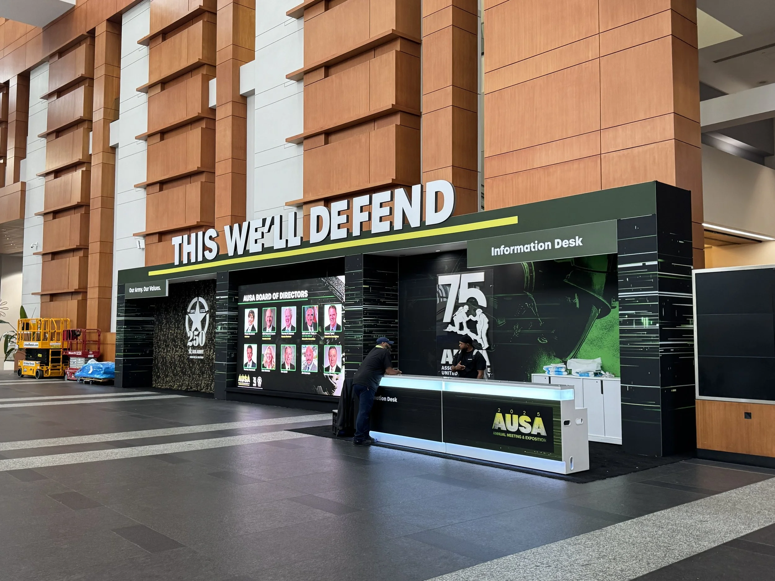 A large indoor space with a high ceiling, featuring an arching brick wall and an information desk that reads 'THIS WE'LL DEFEND.' The desk has various digital displays and signs related to a military or defense conference. Two men are standing at the