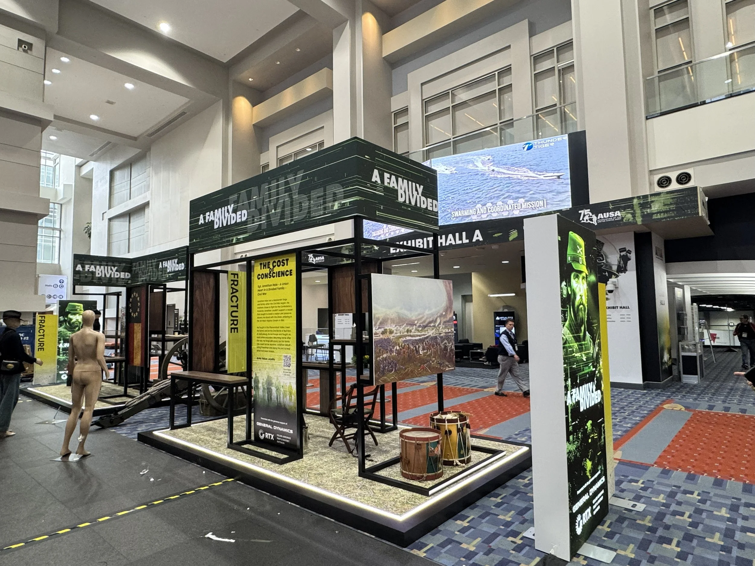 Exhibit booth at a convention with display panels titled 'A Family Divided' and 'The Cost of Conscience', featuring images of military aircraft, historical sites, and a person in military gear. There are attendees walking around, some looking at the 