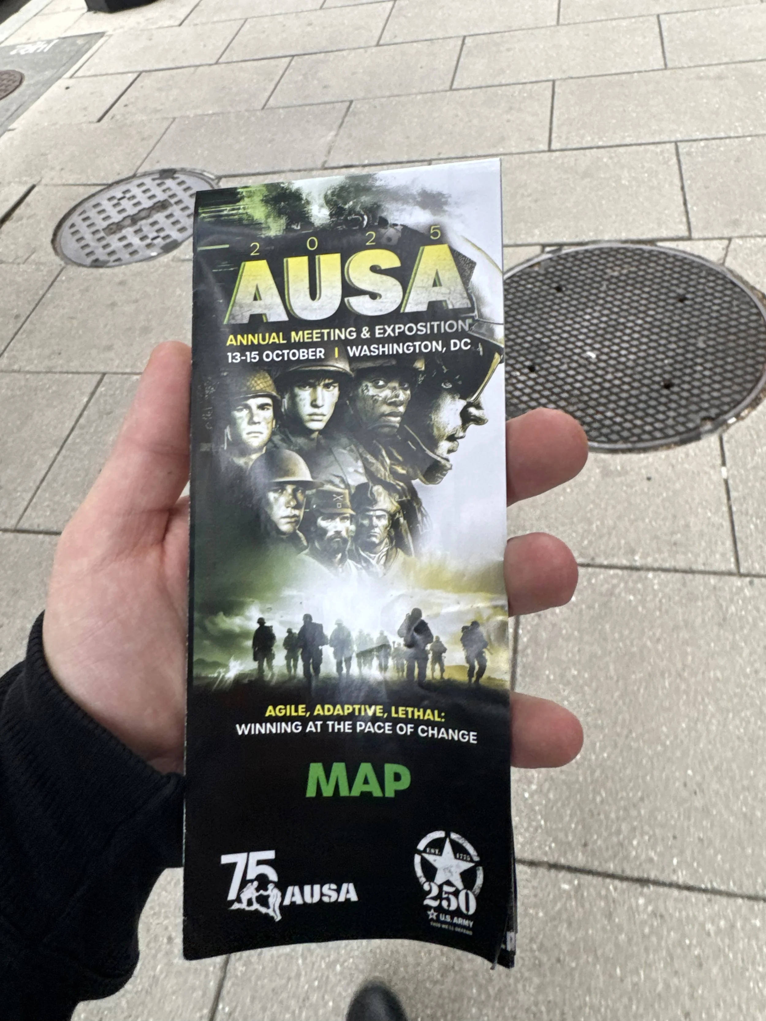 A person holding a brochure for the 2025 AUSA Annual Meeting & Exposition in Washington, DC, with soldiers in a combat scene and a green military-themed design.