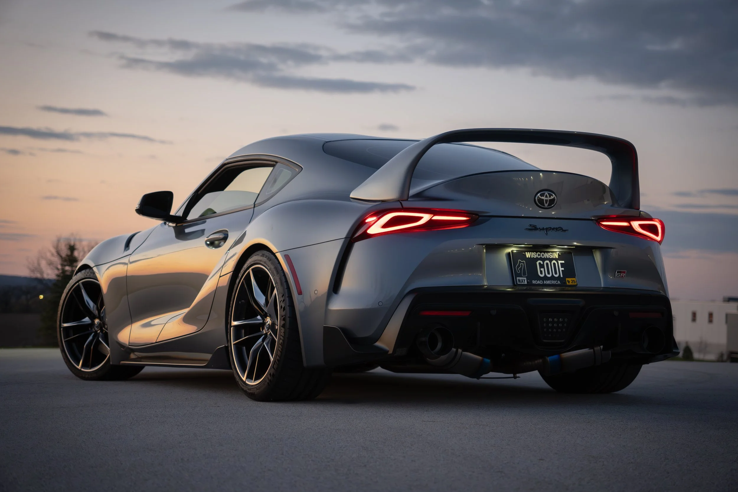 A silver Toyota Supra sports car with a custom Wisconsin license plate reading 'GOOF' parked outdoors during sunset, featuring sleek design and aerodynamic features.