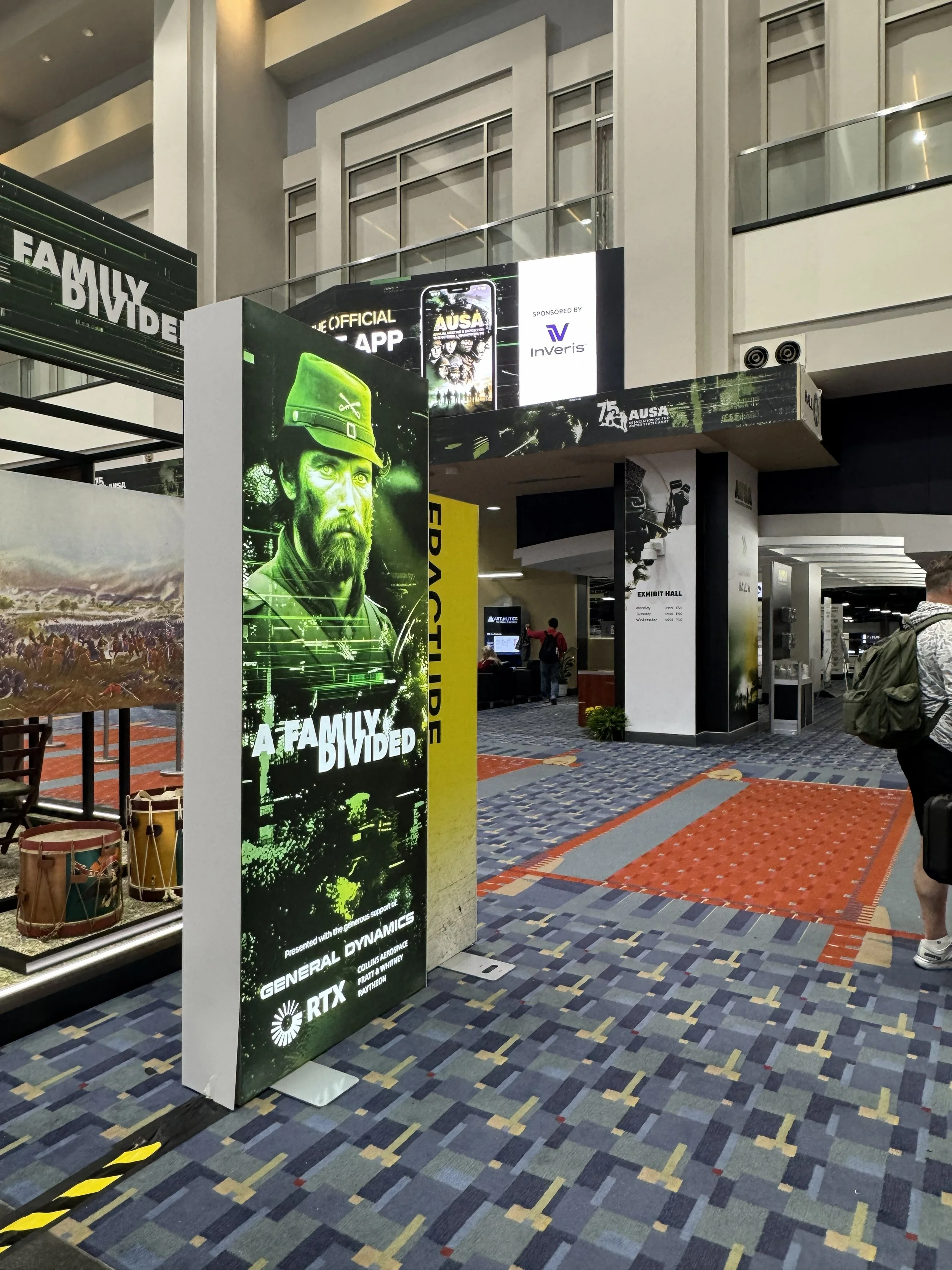 Indoor convention center with promotional signs for a video game, including a large poster of a soldier with a green helmet and the text 'A FAMILY DIVIDED.' There are digital screens displaying game-related content, and a walkway with patterned carpe