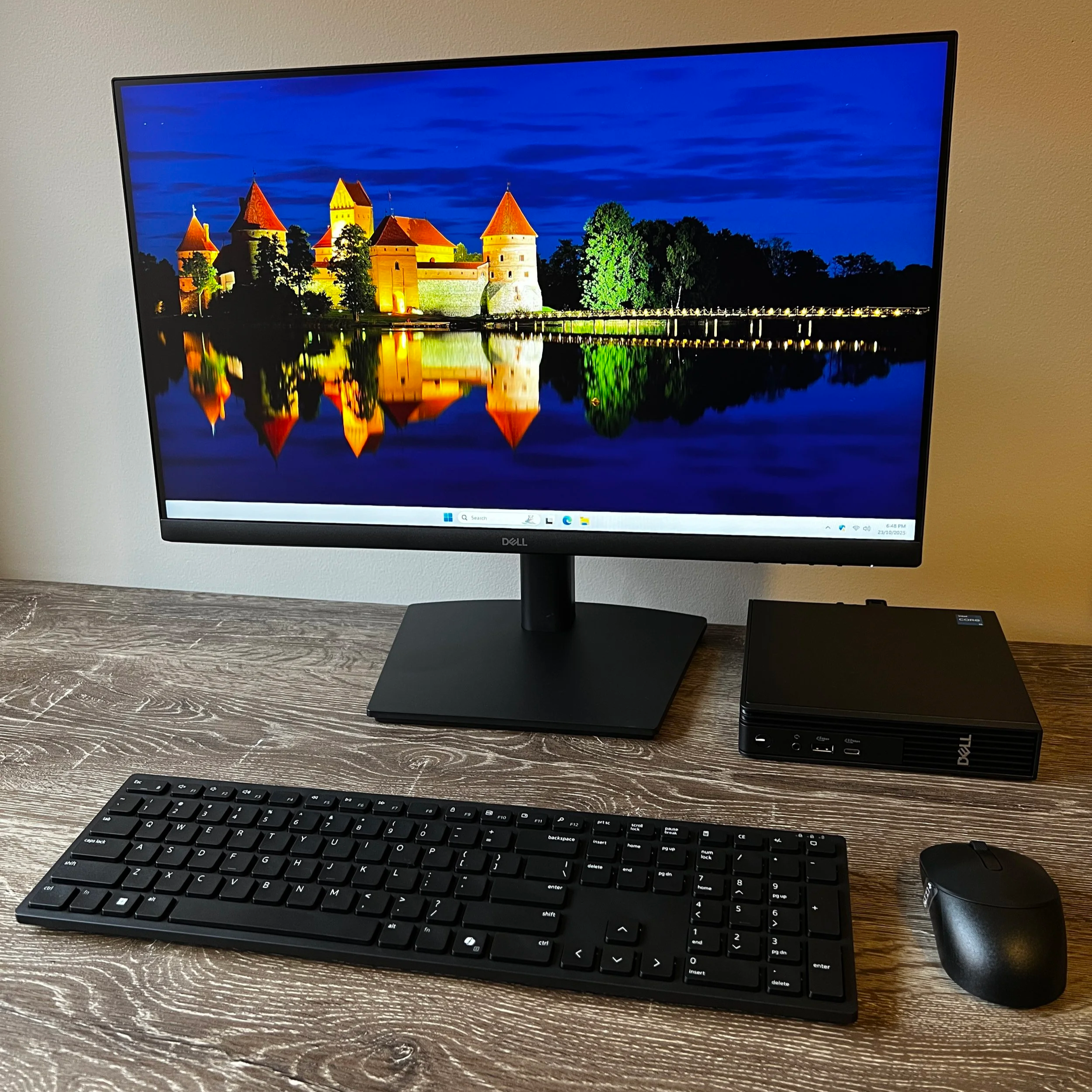 A new Dell desktop setup with a Dell monitor displaying a night view of a castle reflected in water, a black keyboard, and a black mouse on a wooden desk.