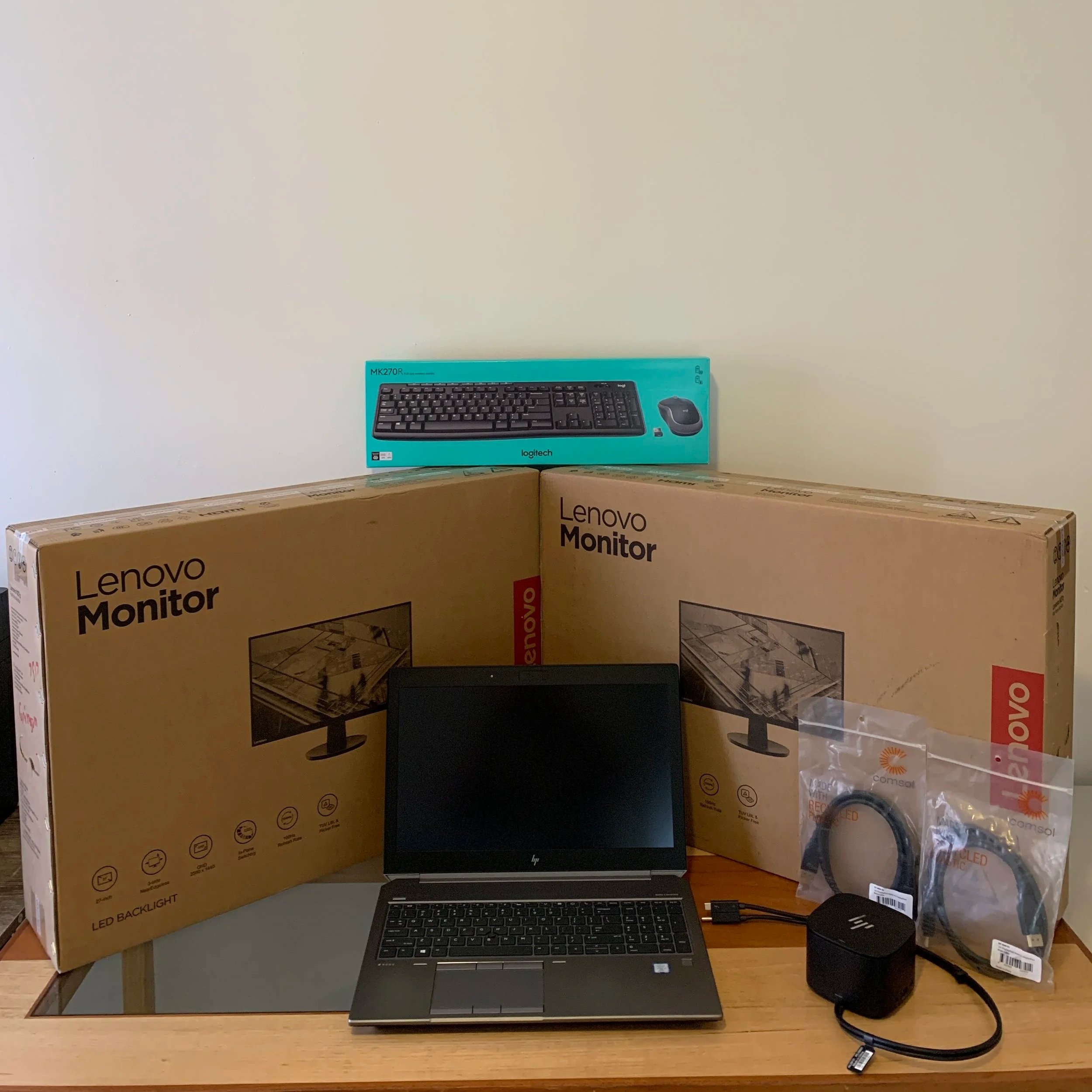 A new HP laptop, two Lenovo monitors in boxes, a Logitech keyboard and mouse, and a power adapter with cables on a wooden desk against a plain wall.
