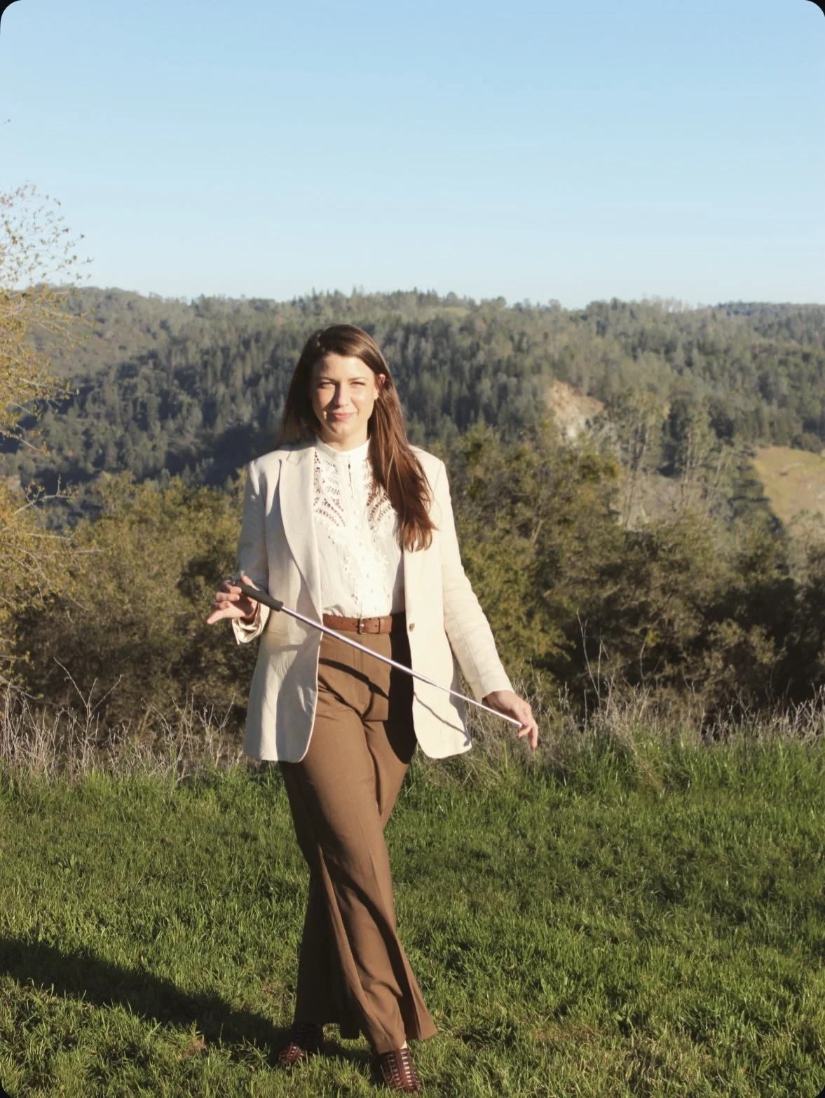 A woman with long brown hair dressed in a cream blazer, cooking shirt, and brown pants, holding a brainspotting pointer, standing outdoors with green grass and trees, and a hill in the background under a blue sky.