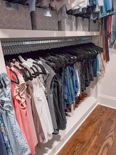 Clothes hanging on black hangers on a white clothing rack with a shelf above and a wooden floor.