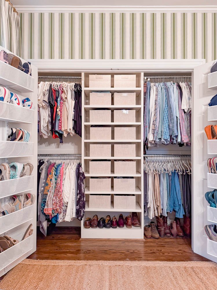 Organized walk-in closet with shelves of shoes, hanging clothes, and baskets, set against striped wallpaper and a wooden floor.