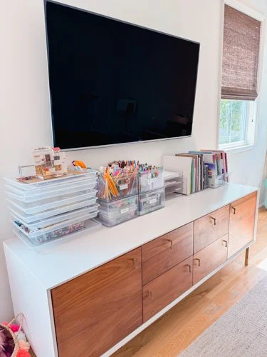A white sideboard with wooden drawers holding organized craft supplies, positioned beneath a wall-mounted flat-screen TV and next to a window with a brown shade.