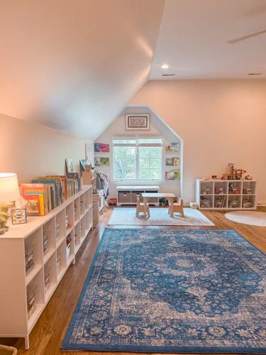 Children's playroom with white bookshelves, a blue patterned rug, and natural light from a window.